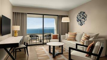 A hotel room with a flat-screen TV, a dining table, a sofa, a coffee table, and a view of the ocean.
