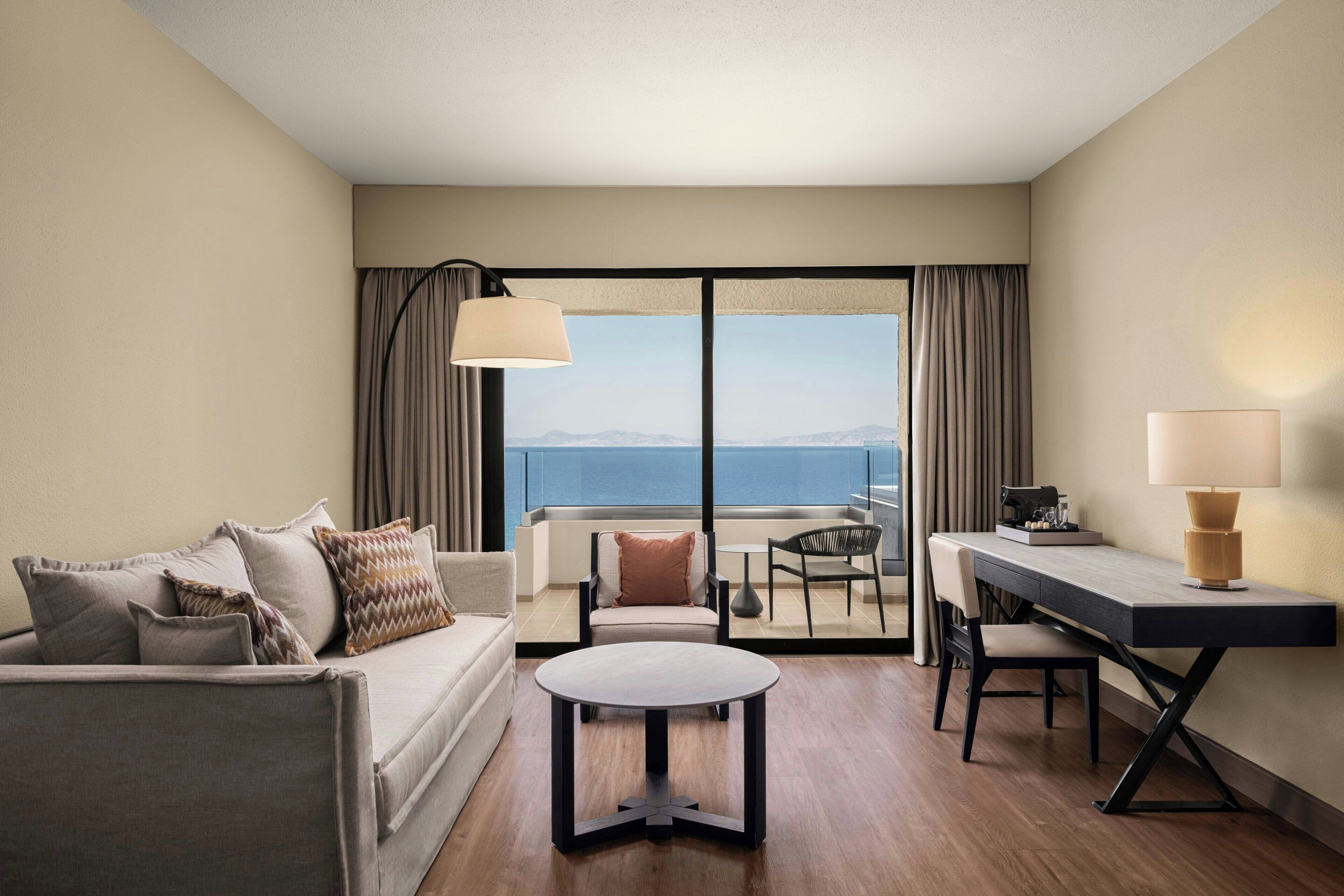 A hotel room with a sofa, a round table, a desk, and a view of the sea.