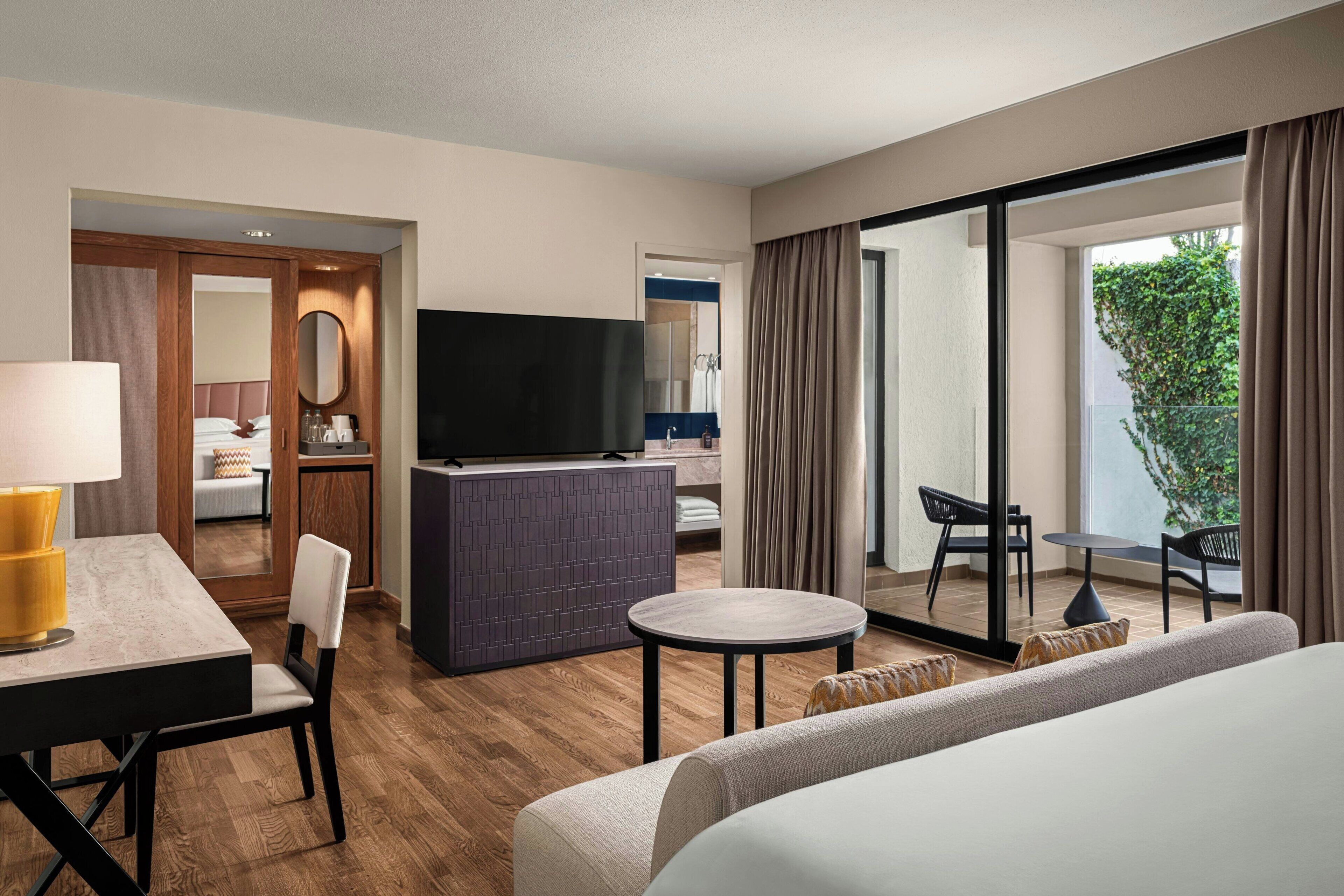 A hotel room with a bed, a TV, a desk with a lamp, a small round table, and a balcony with a view of greenery.