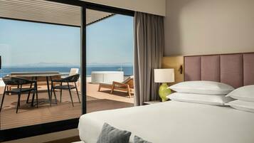 Une chambre d’hôtel avec un grand lit, une table de chevet, une lampe, une fenêtre donnant sur la mer et un balcon avec des sièges.