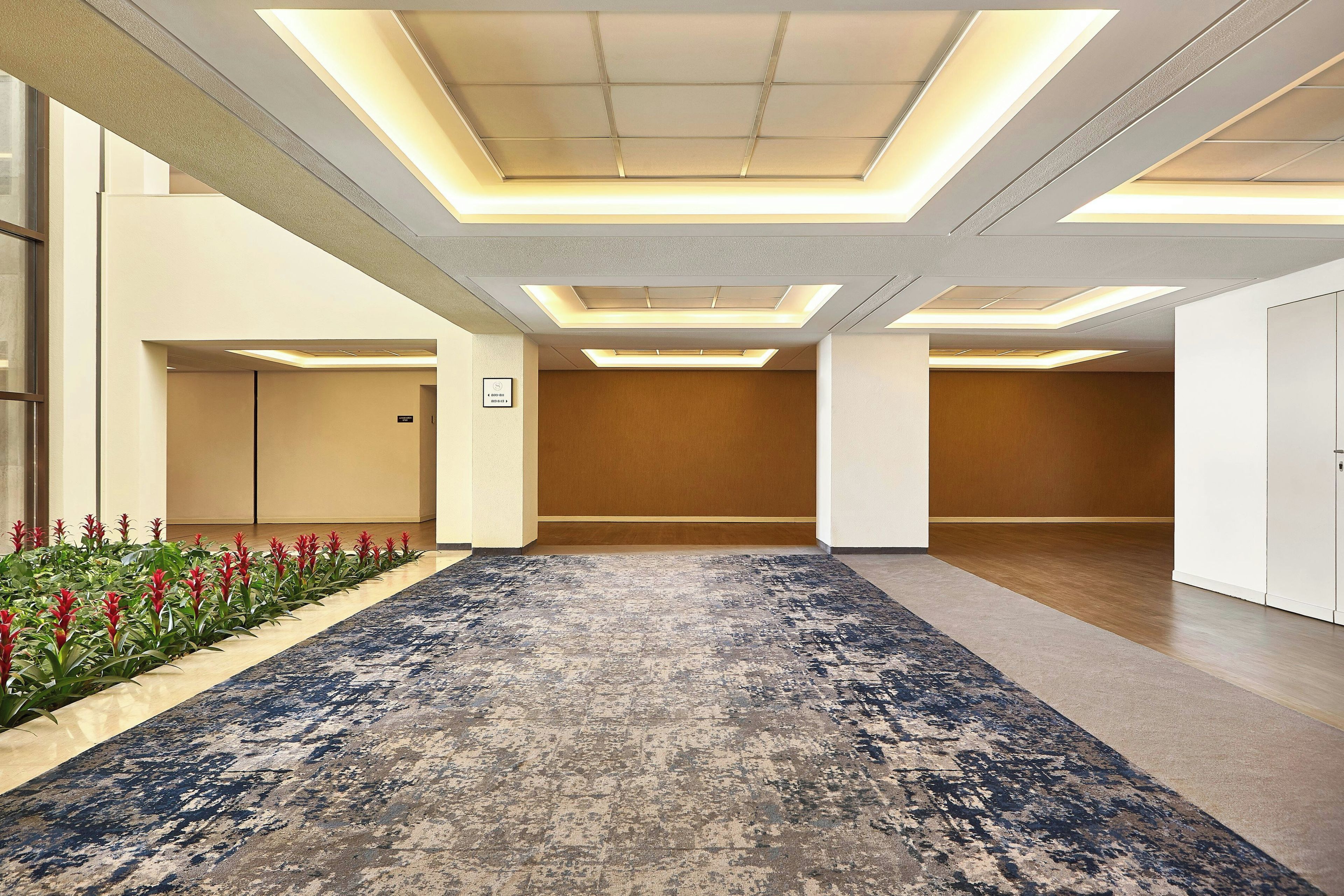 A hotel corridor with a patterned carpet, recessed ceiling lights, and a wooden panel wall.
