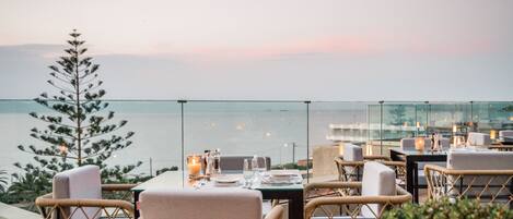 A rooftop dining area with wicker chairs, tables set for dining, and a view of the sea.