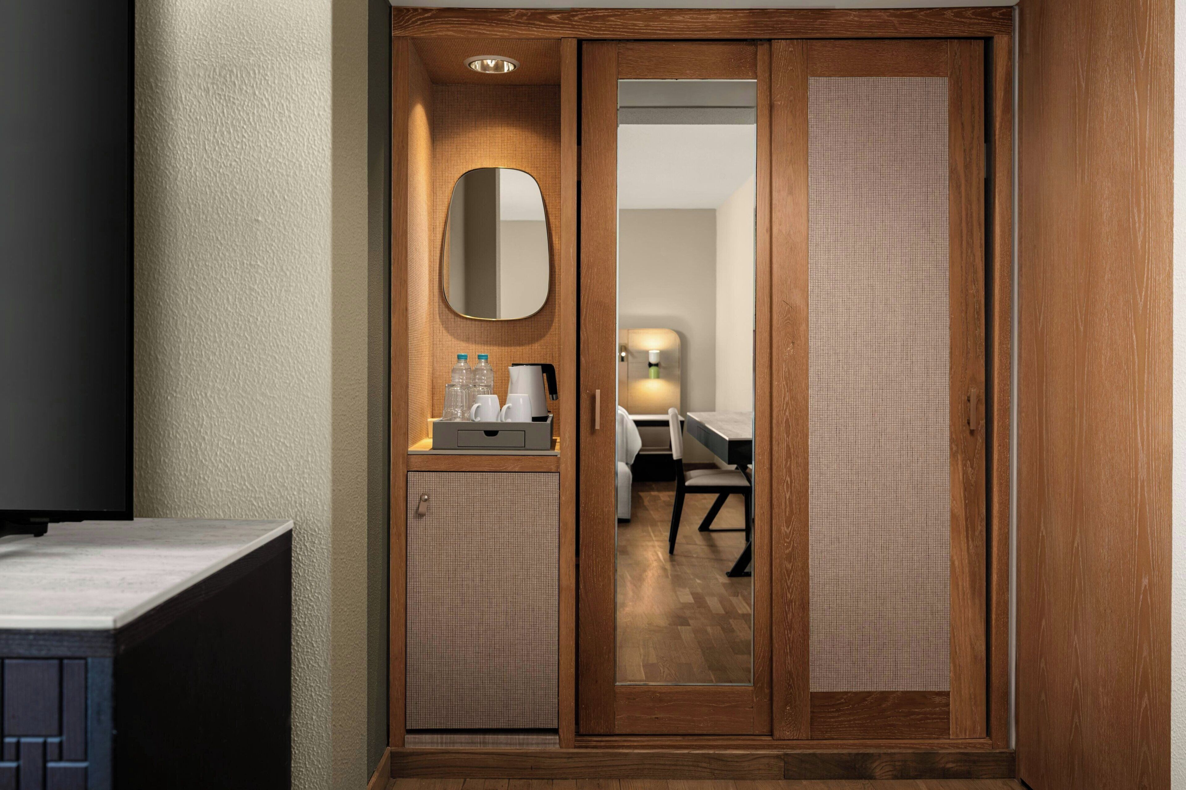 A hotel room with a wooden wardrobe, a small kitchenette with a coffee maker, and a mirror.