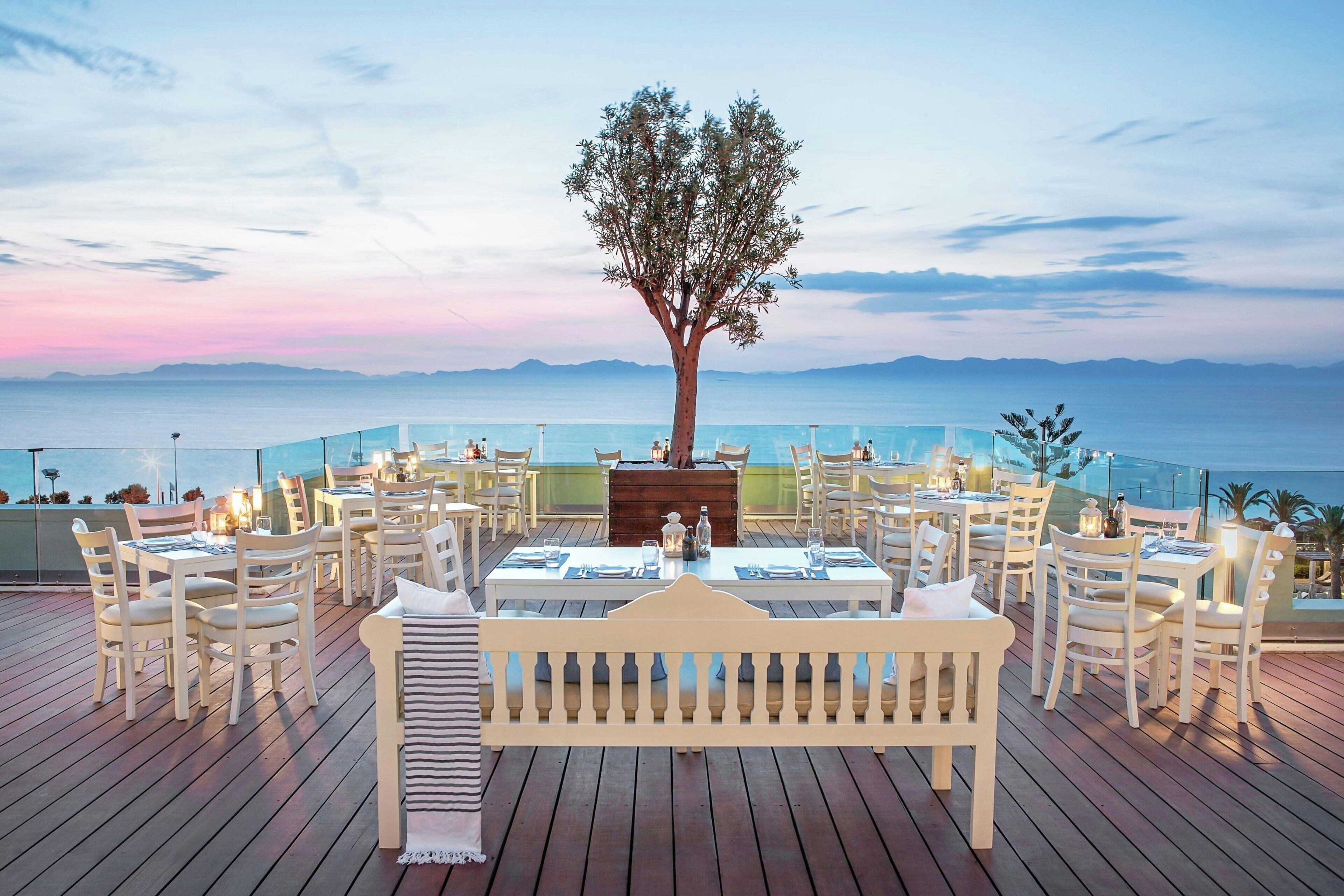 Un área de comedor al aire libre con mesas y sillas, una terraza de madera y vista al mar.