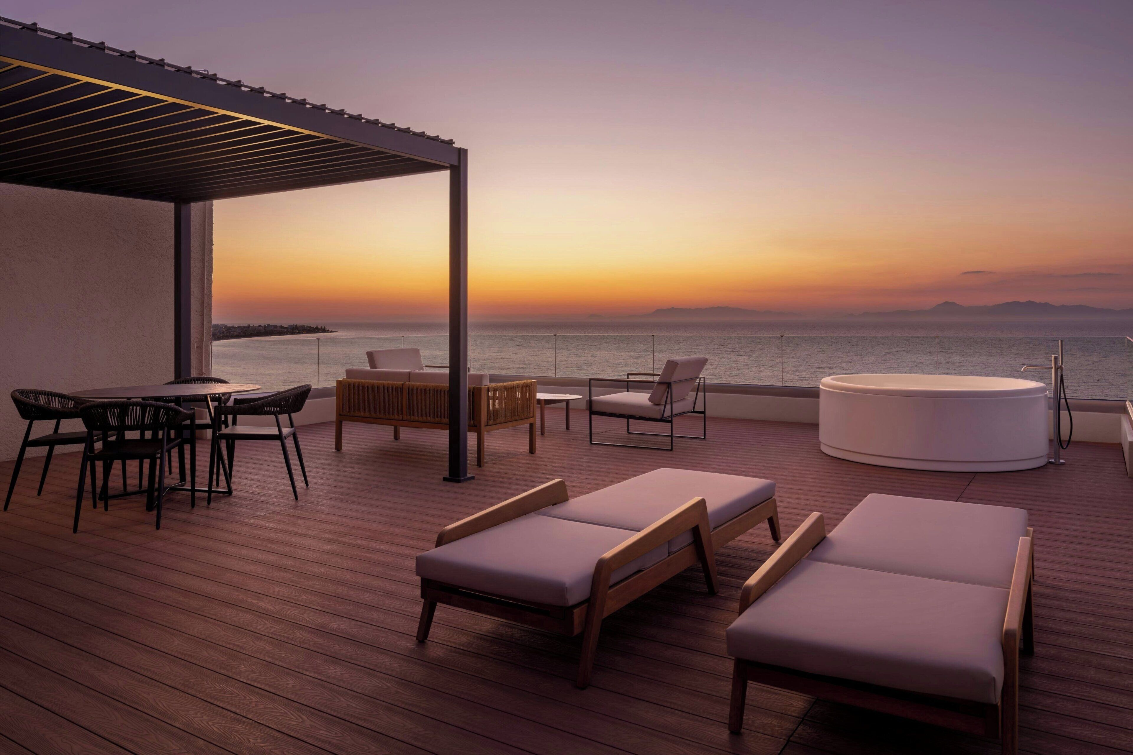 Terraza con sillones de descanso, una mesa redonda y un sofá, con vista a un cuerpo de agua al atardecer.