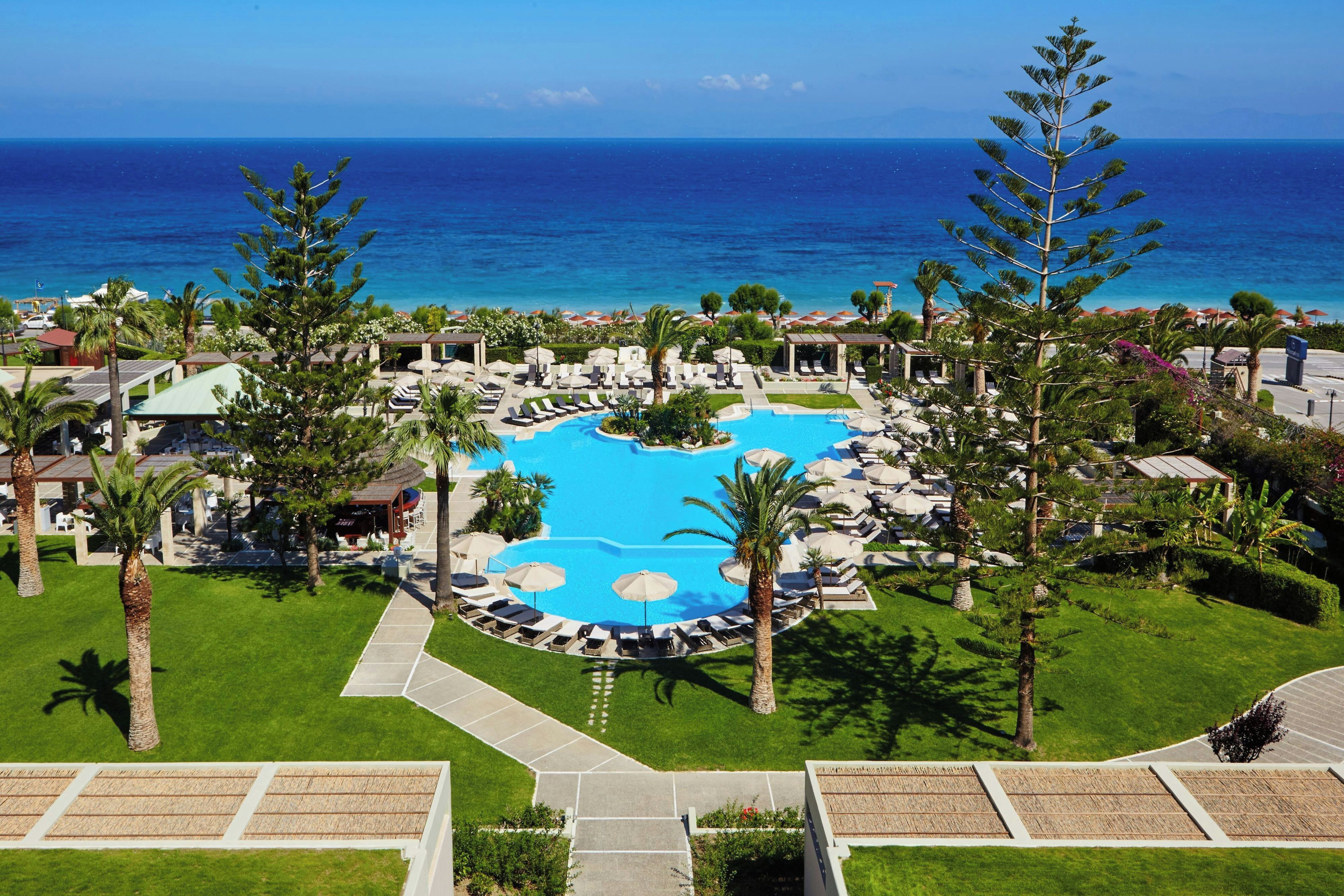 A resort with a central swimming pool surrounded by lounge areas and palm trees, overlooking a beach.