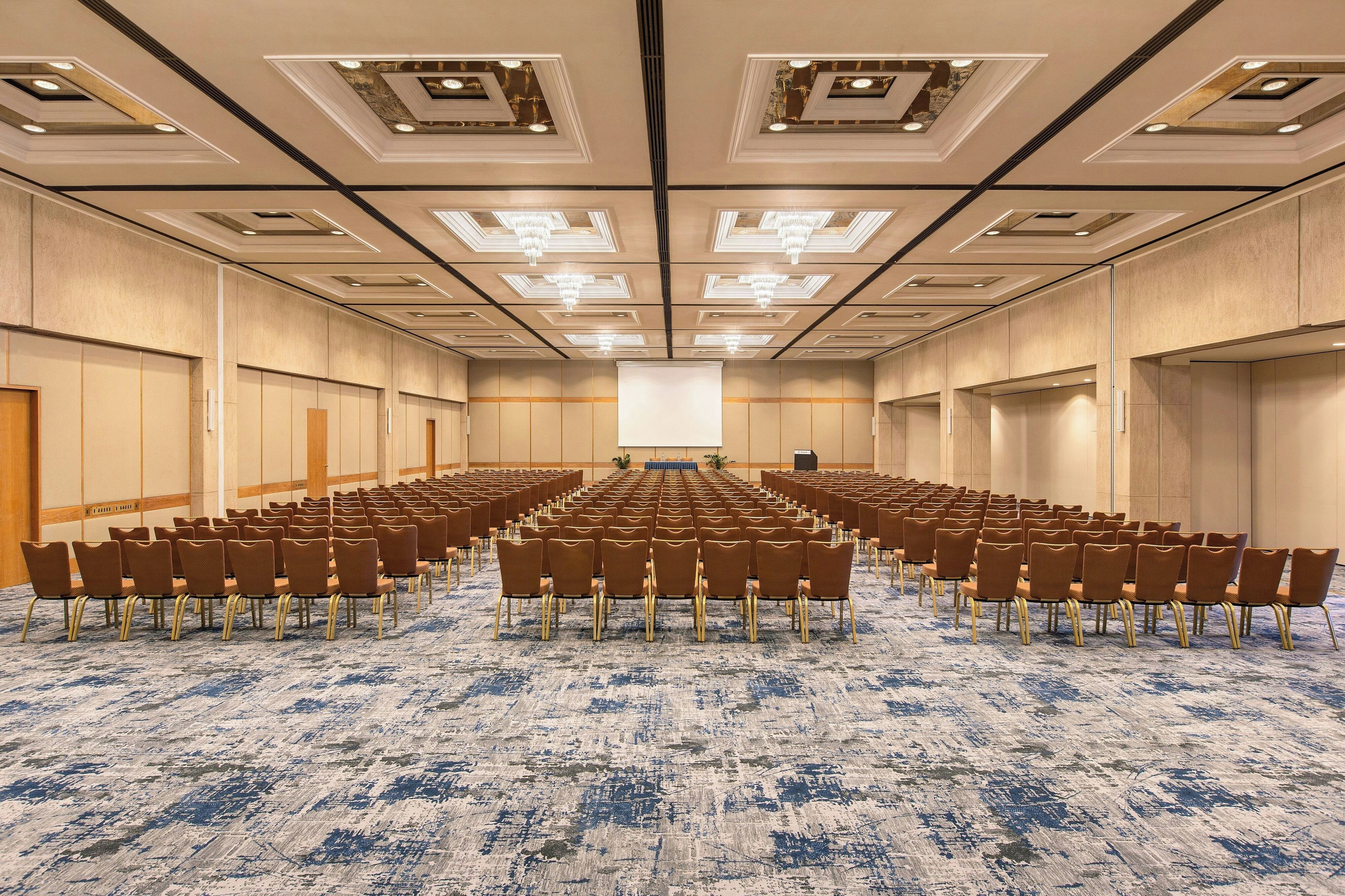 A conference room with rows of chairs, a stage, and a large screen.