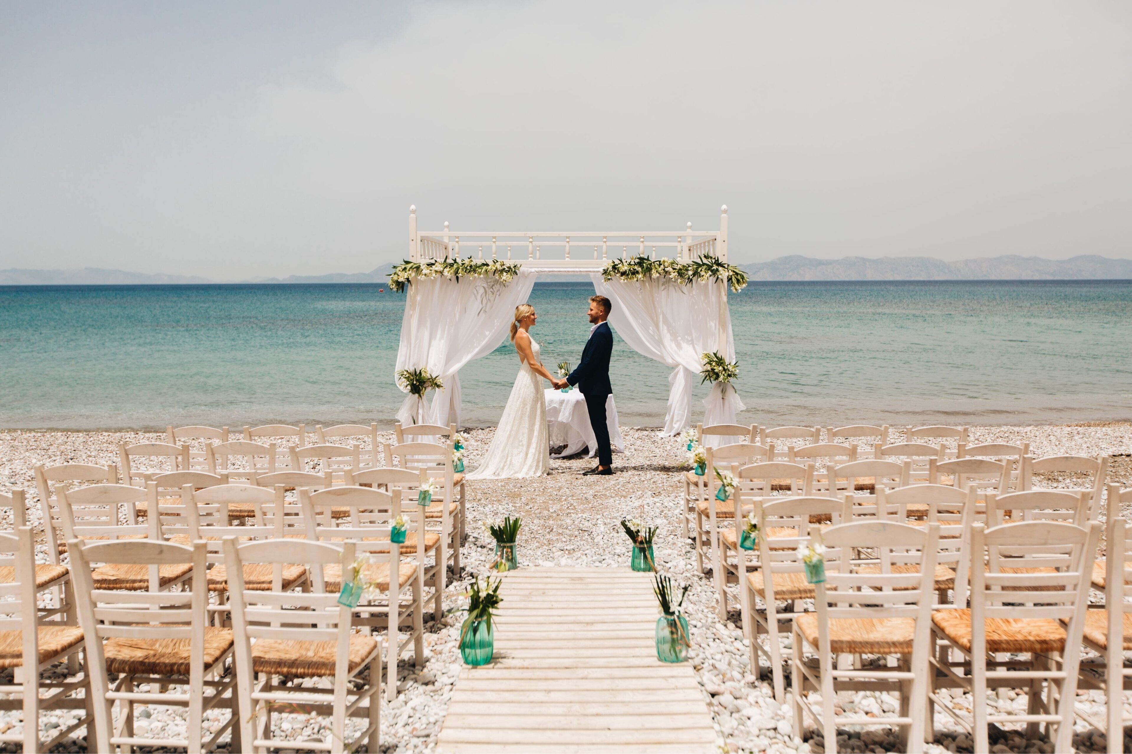 Ceremonia de boda en la playa, con un arco blanco y sillas dispuestas para los invitados.