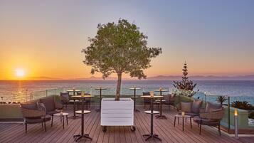 A deck with outdoor seating, a tree, and a view of the ocean at sunset.