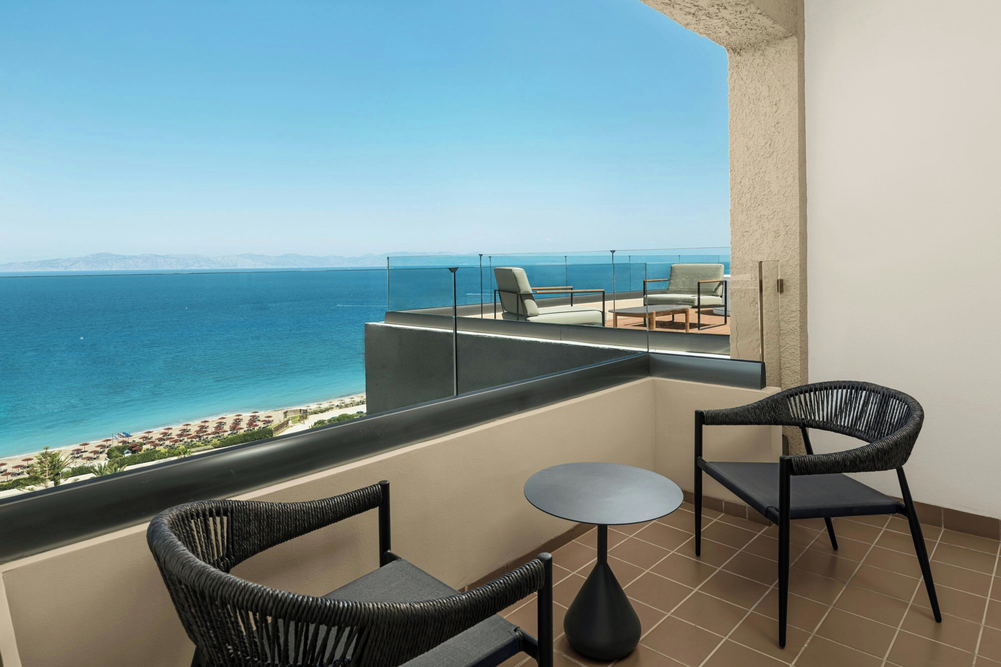 A balcony with two wicker chairs and a small round table, overlooking a beach with umbrellas and a clear blue sea.