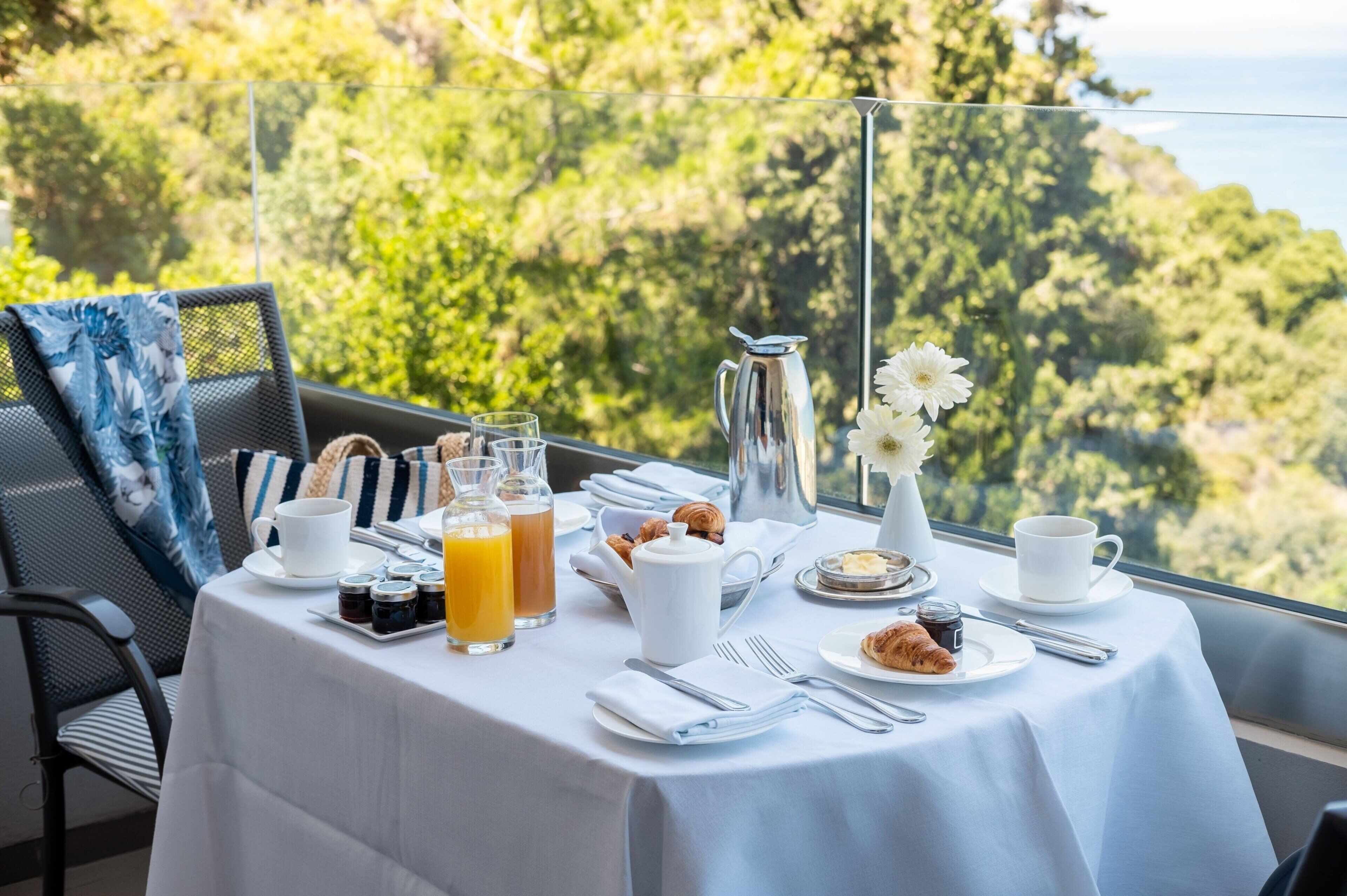 Une table de petit-déjeuner dressée avec des croissants, des pâtisseries, un pichet, des tasses et des verres à jus, donnant sur un paysage verdoyant et luxuriant.