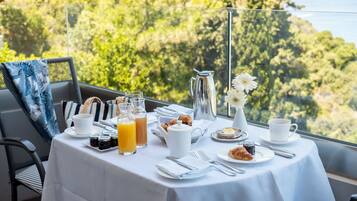 Une table de petit-déjeuner dressée avec des croissants, des pâtisseries, un pichet, des tasses et des verres à jus, donnant sur un paysage verdoyant et luxuriant.