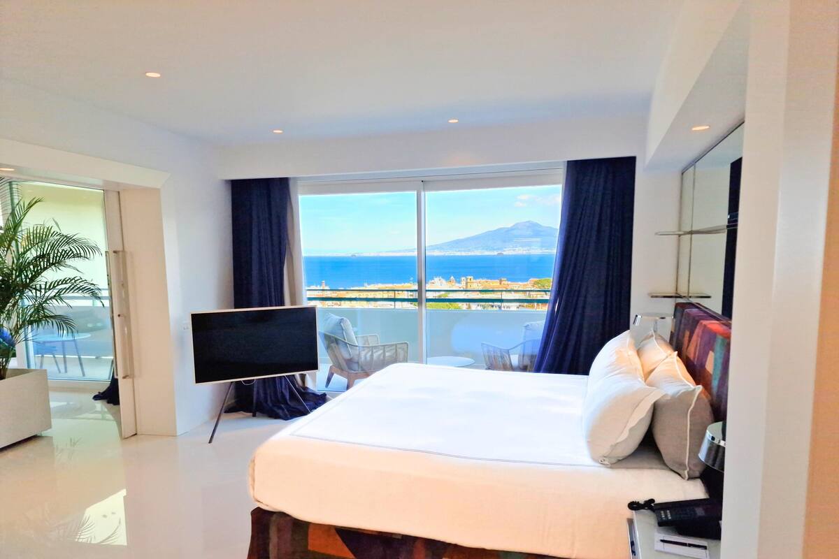 Une chambre d’hôtel moderne dotée d’un grand lit, d’un téléviseur à écran plat, d’un balcon offrant une vue sur la mer et les montagnes, et d’une plante en pot.