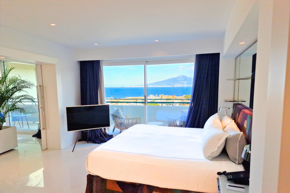 A modern hotel room with a large bed, a flat-screen TV, a balcony with a view of the sea and a mountain, and a potted plant.