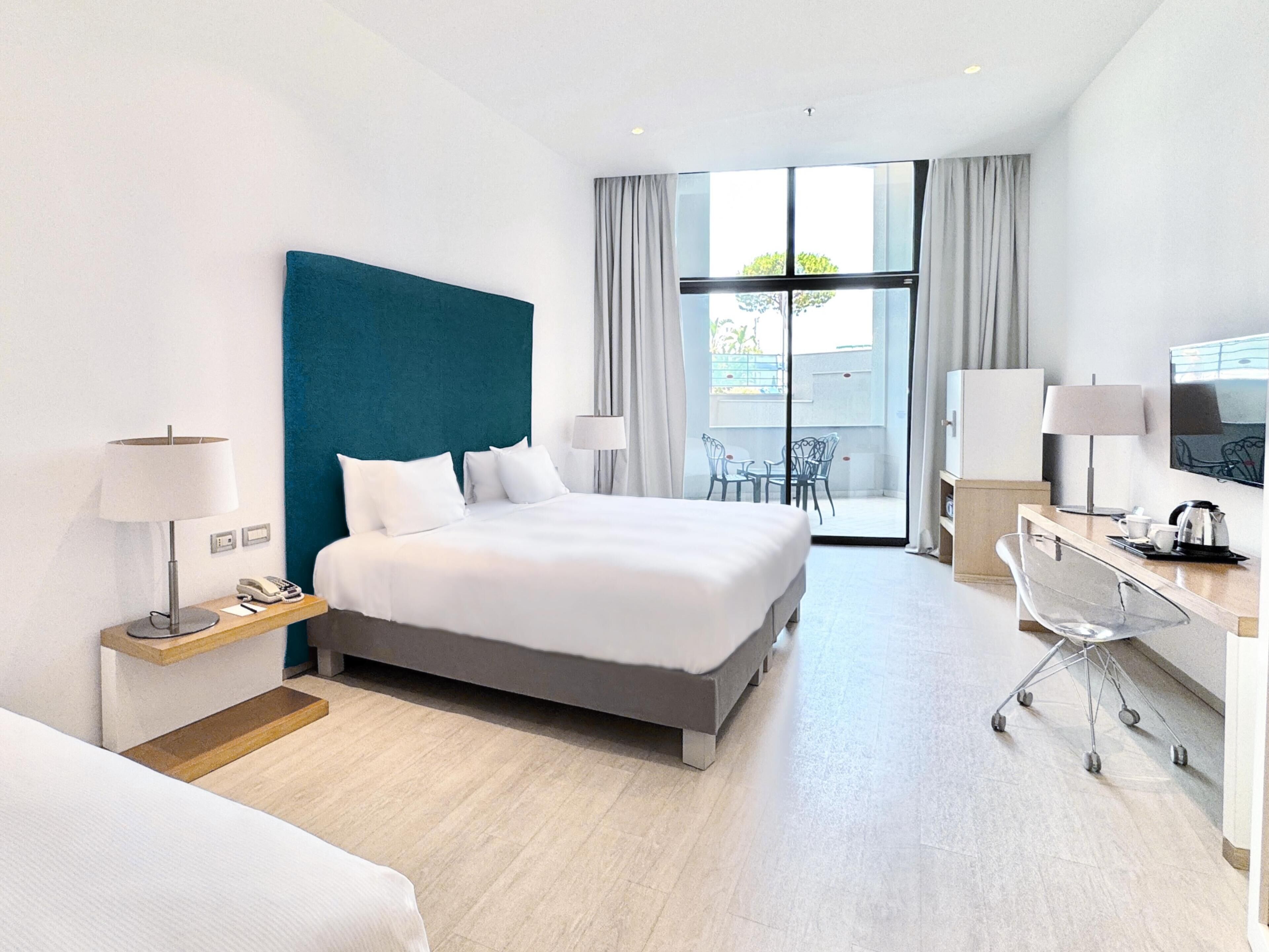 A modern hotel room with a large bed, a desk, a chair, and a balcony with a table and chairs.