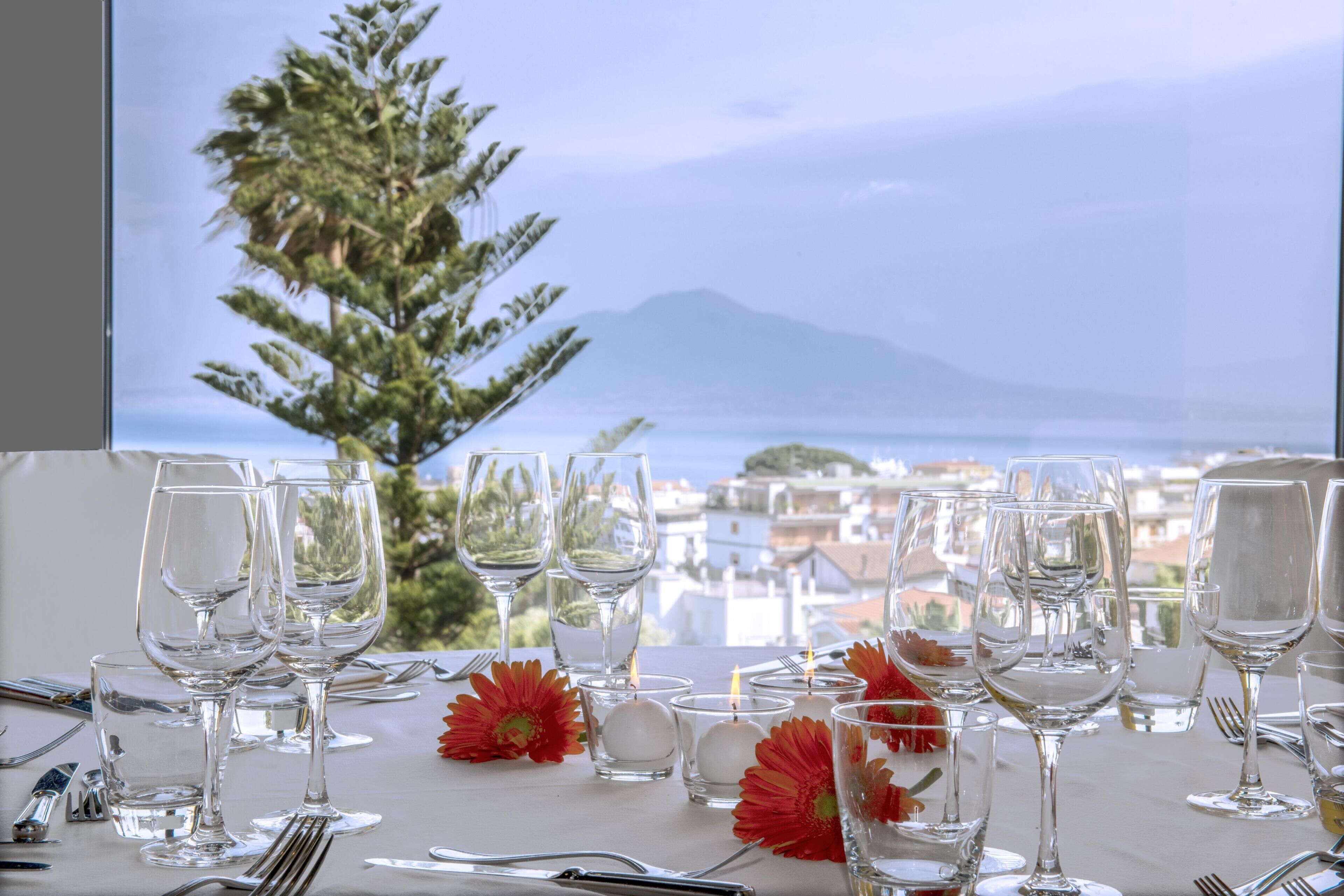 A table set for dining with clear glassware and candles, overlooking a coastal town and a mountain.