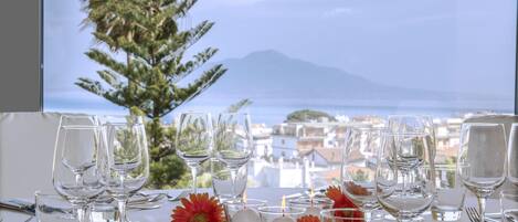A table set for dining with clear glassware and candles, overlooking a coastal town and a mountain.