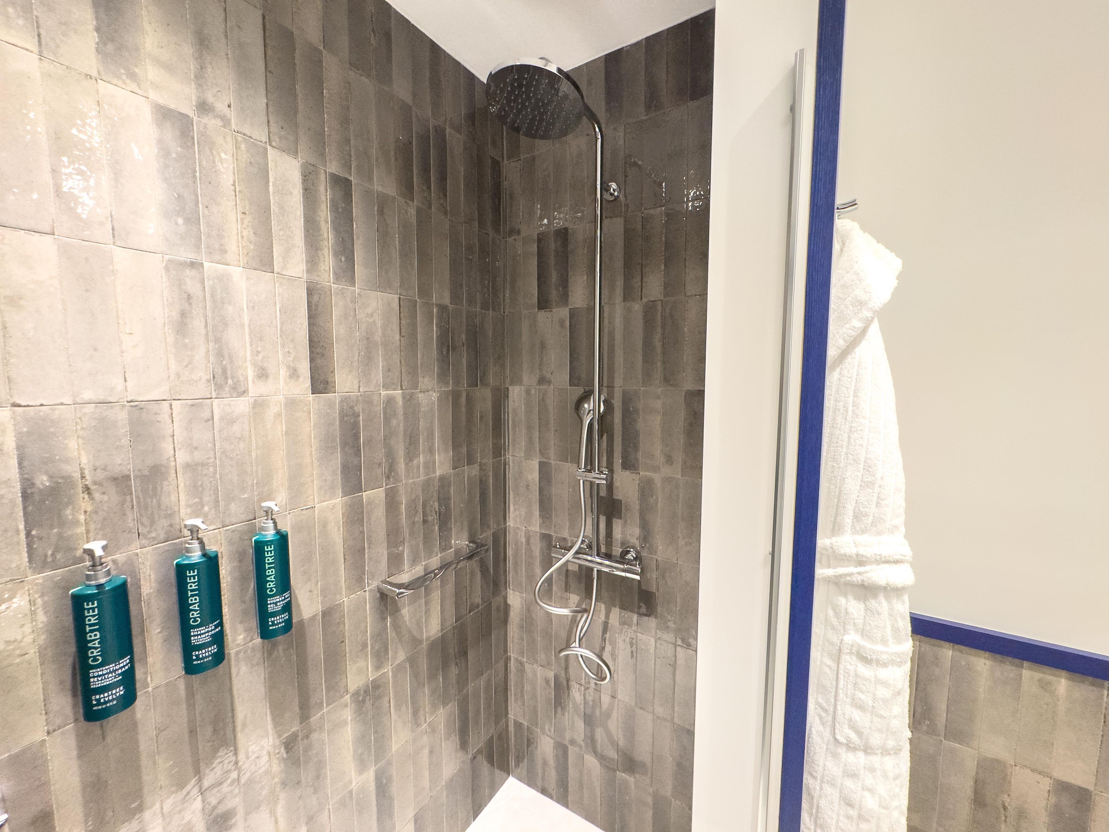 Un baño con ducha, paredes de azulejos y tres botellas de champú Crabtree.