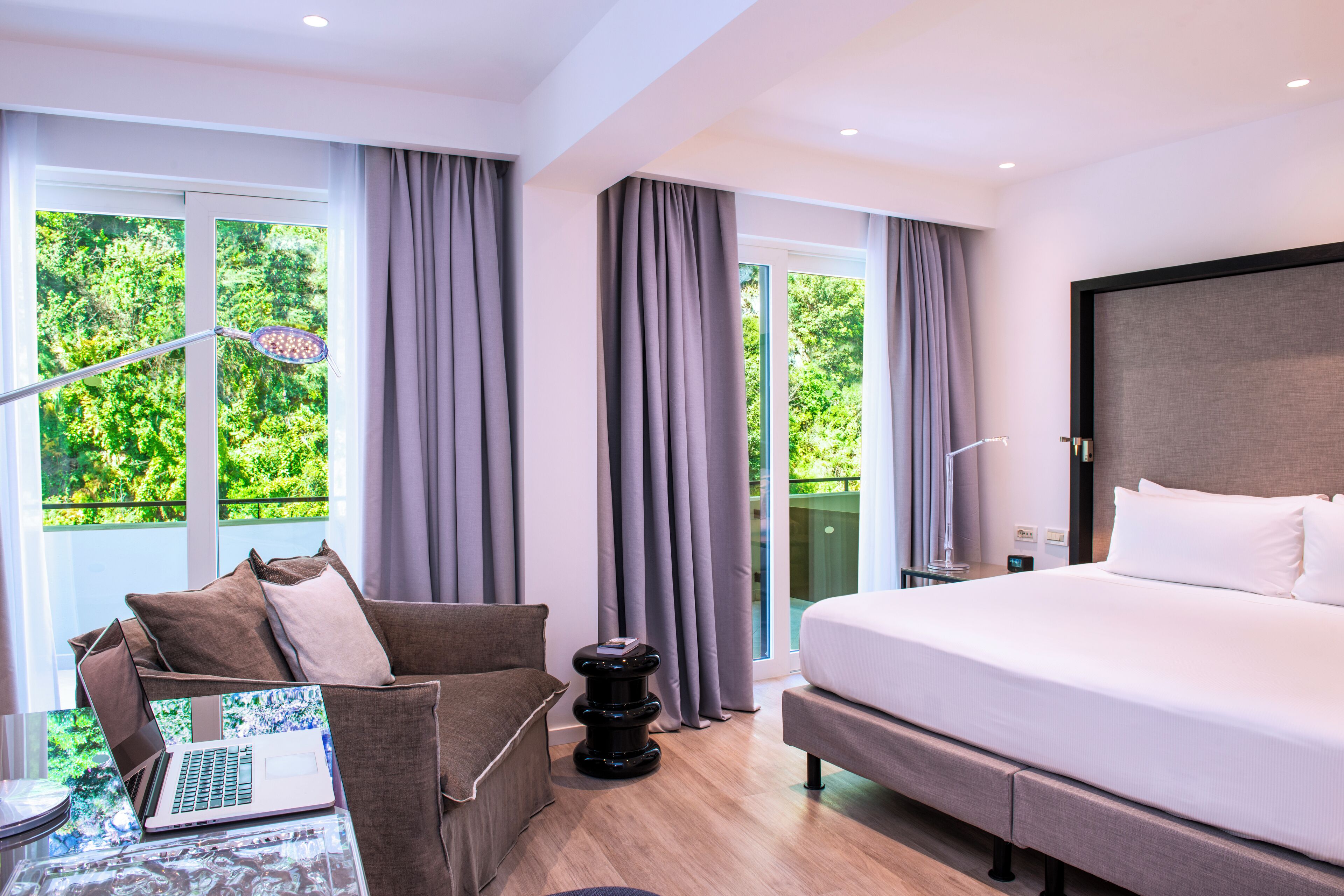 A modern hotel room with a large bed, a sitting area with a laptop, and large windows with curtains.