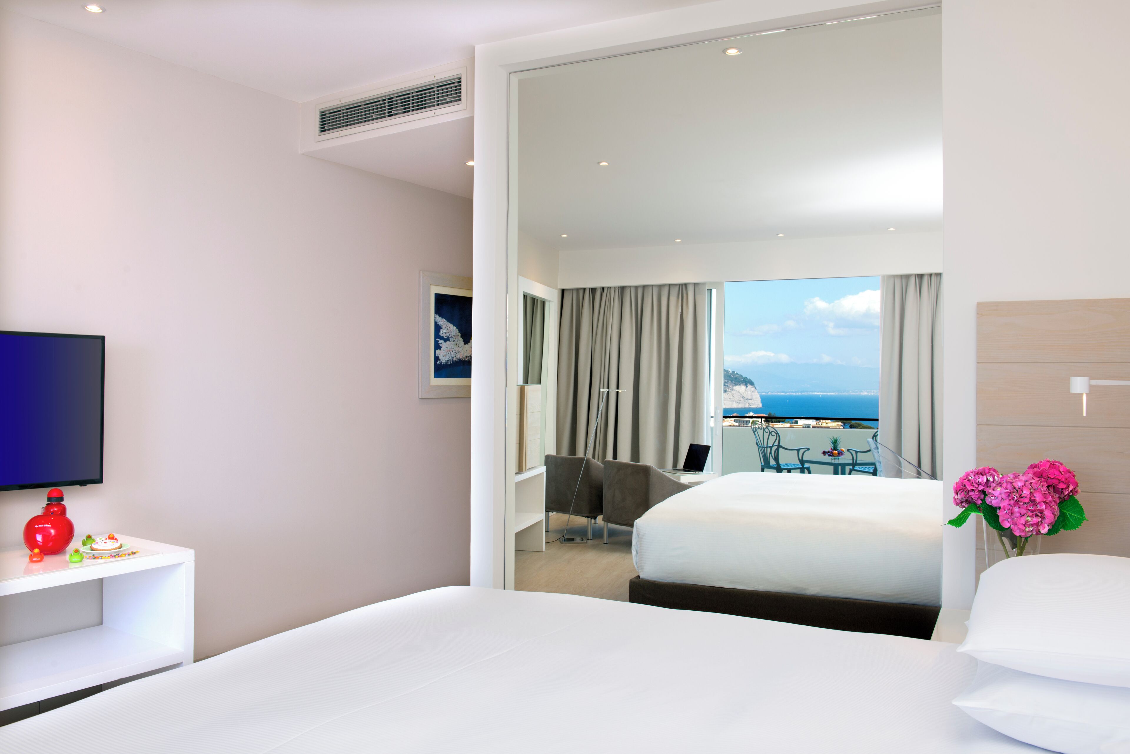 A hotel room with a bed, a TV, a desk with a red lamp, and a view of the sea.
