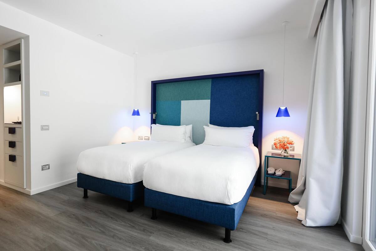 Un lit double avec une tête de lit bleue, deux tables de chevet avec des lampes, une petite table de nuit avec un vase de fleurs et un plancher en bois.