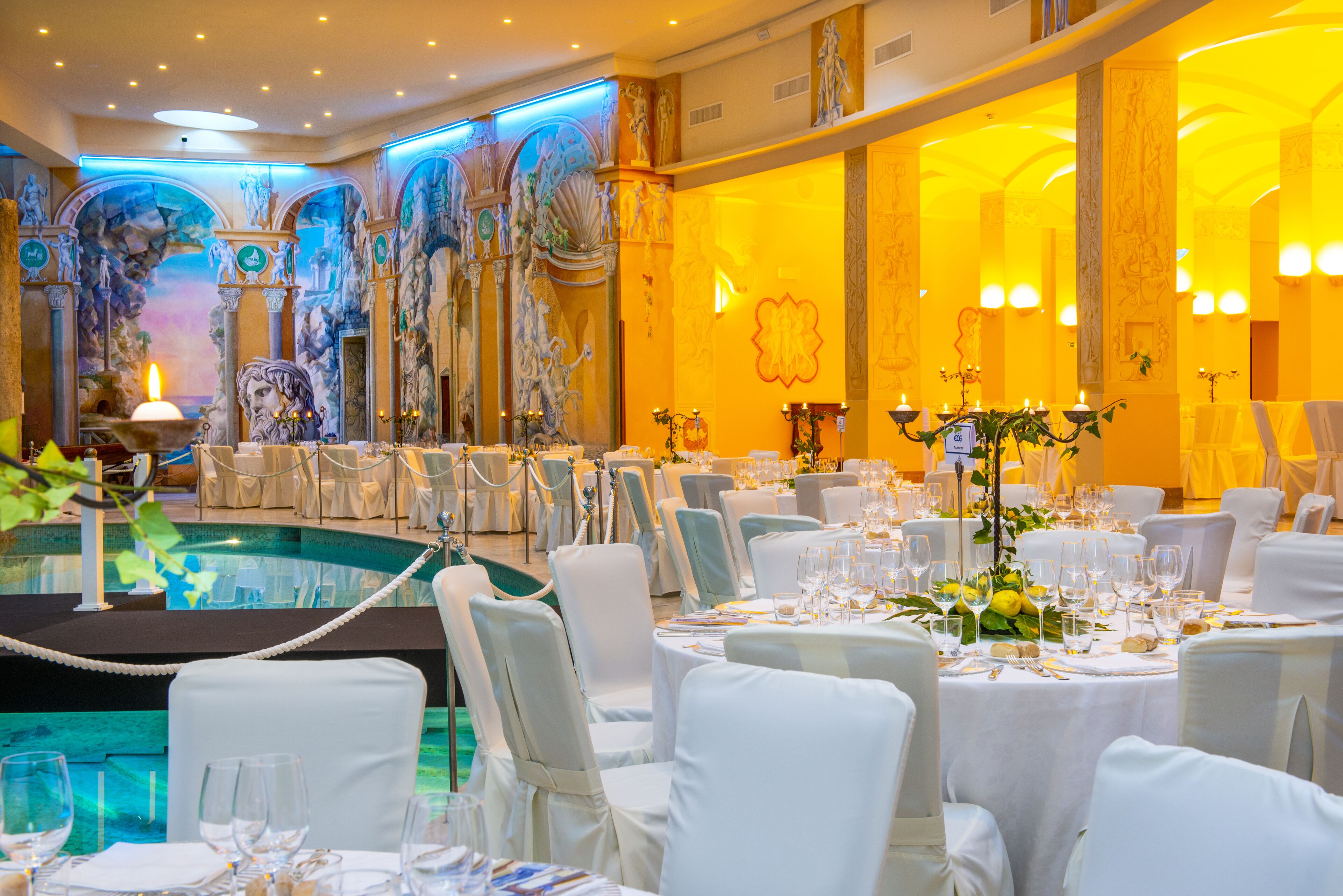 Une salle de banquet avec des tables rondes dressées pour un événement formel, un espace piscine et des œuvres d’art classiques accrochées aux murs.