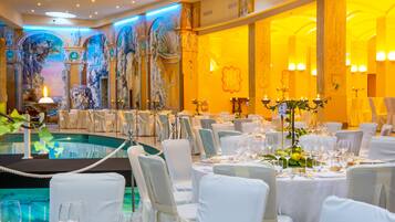 Une salle de banquet avec des tables rondes dressées pour un événement formel, un espace piscine et des œuvres d’art classiques accrochées aux murs.