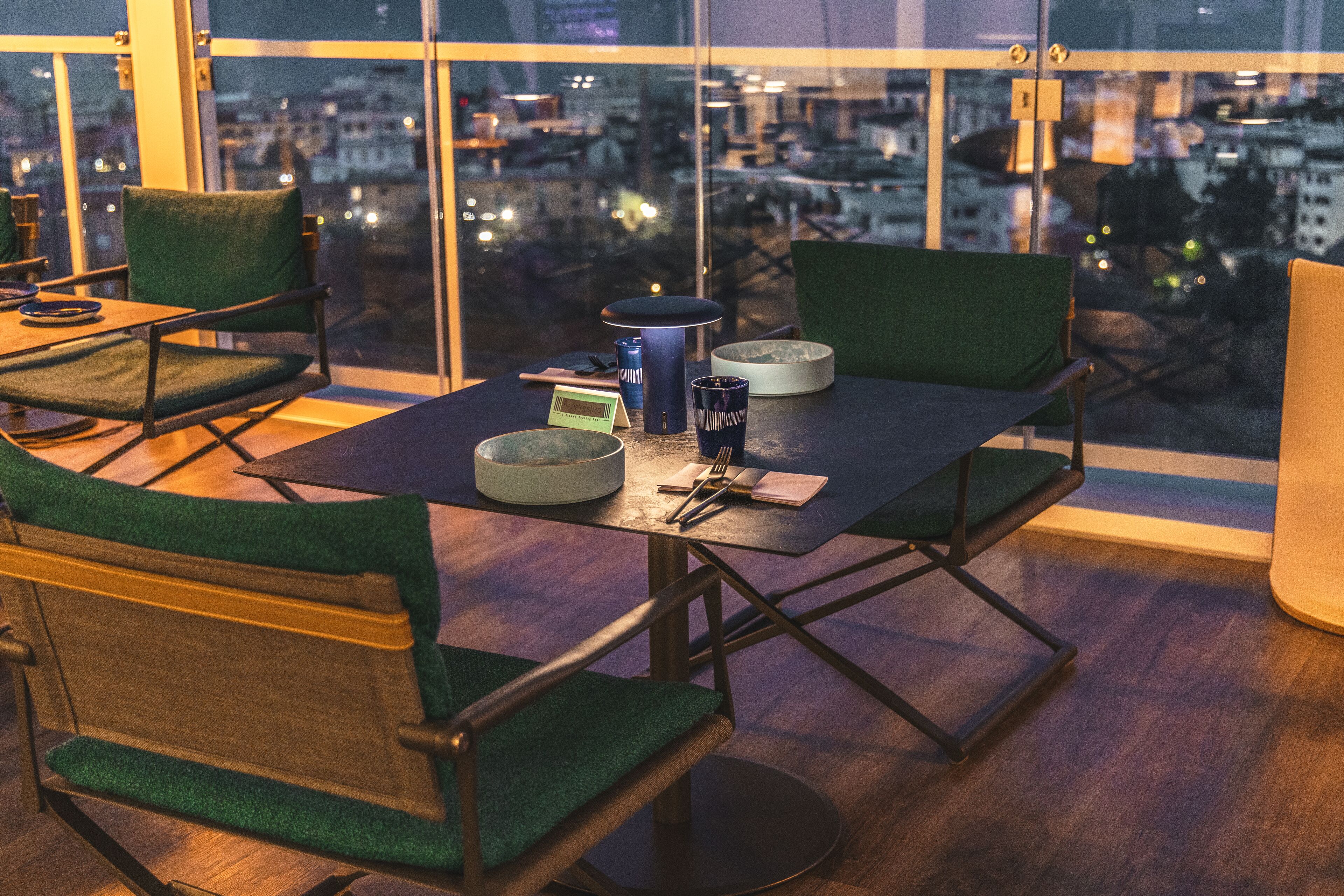 A modern dining area with a table set for two, featuring a city view through large windows.