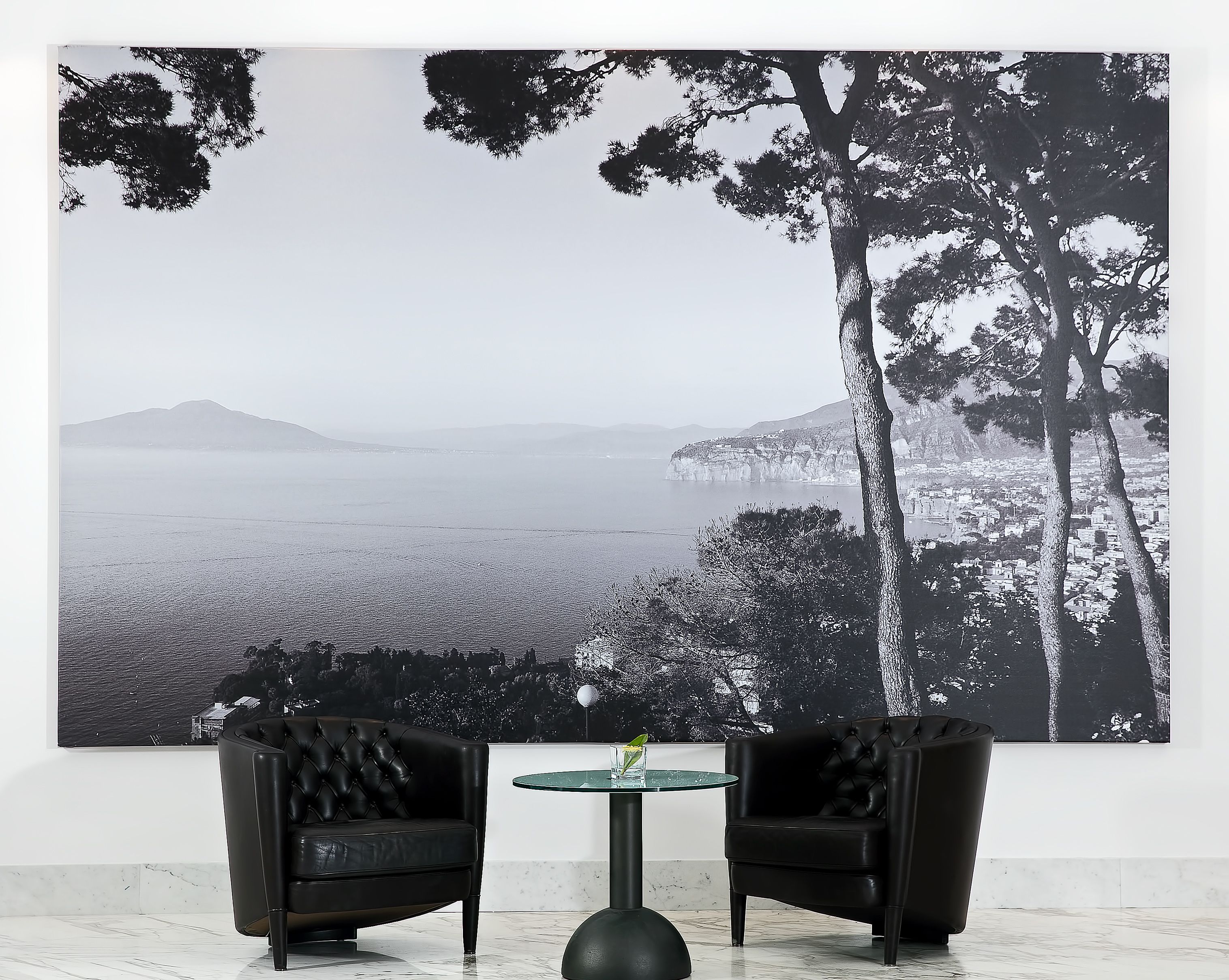 A room with a large black and white landscape painting, two black leather chairs, a glass table, and a marble floor.
