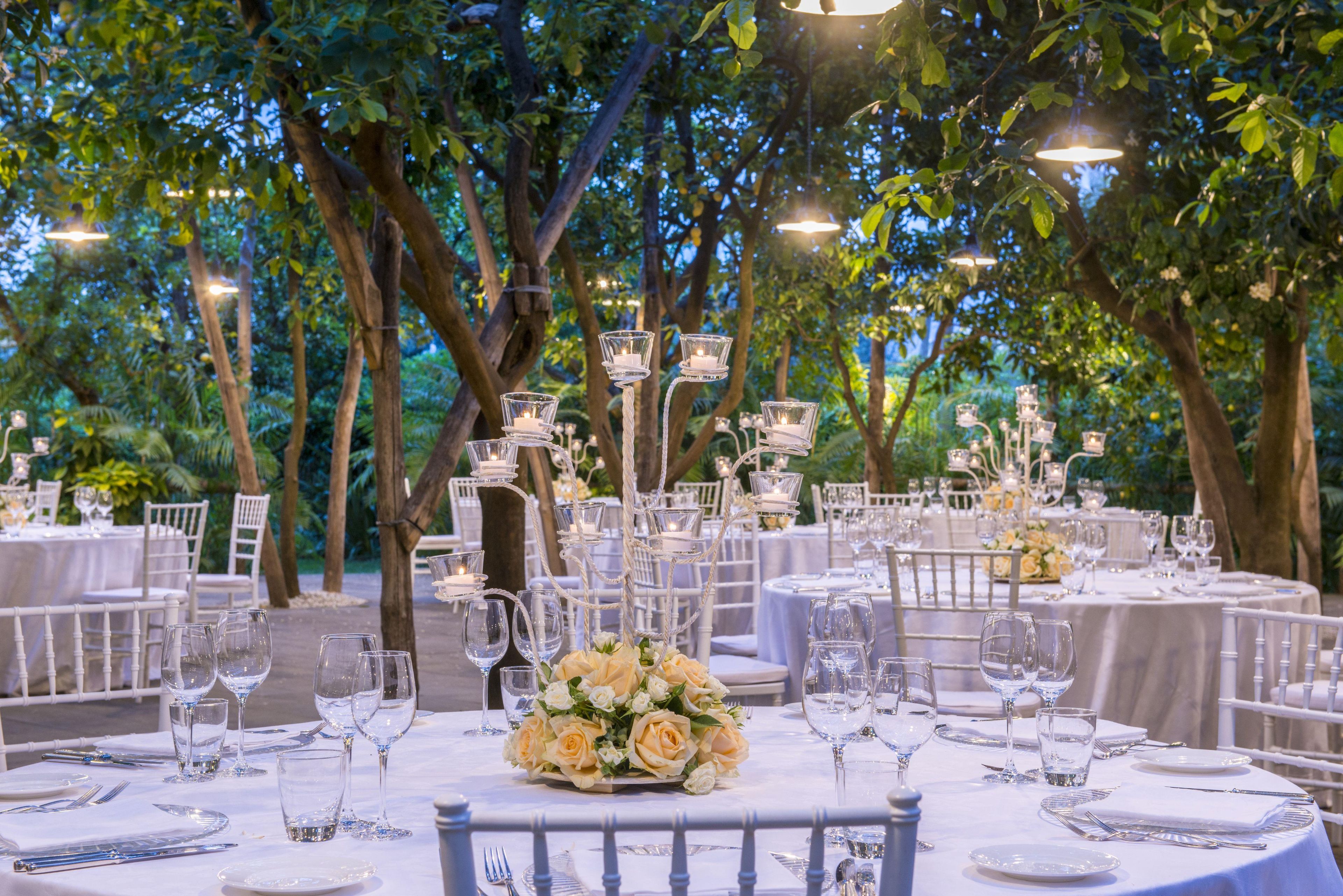 An outdoor dining area with round tables set for a formal event, surrounded by trees and string lights.