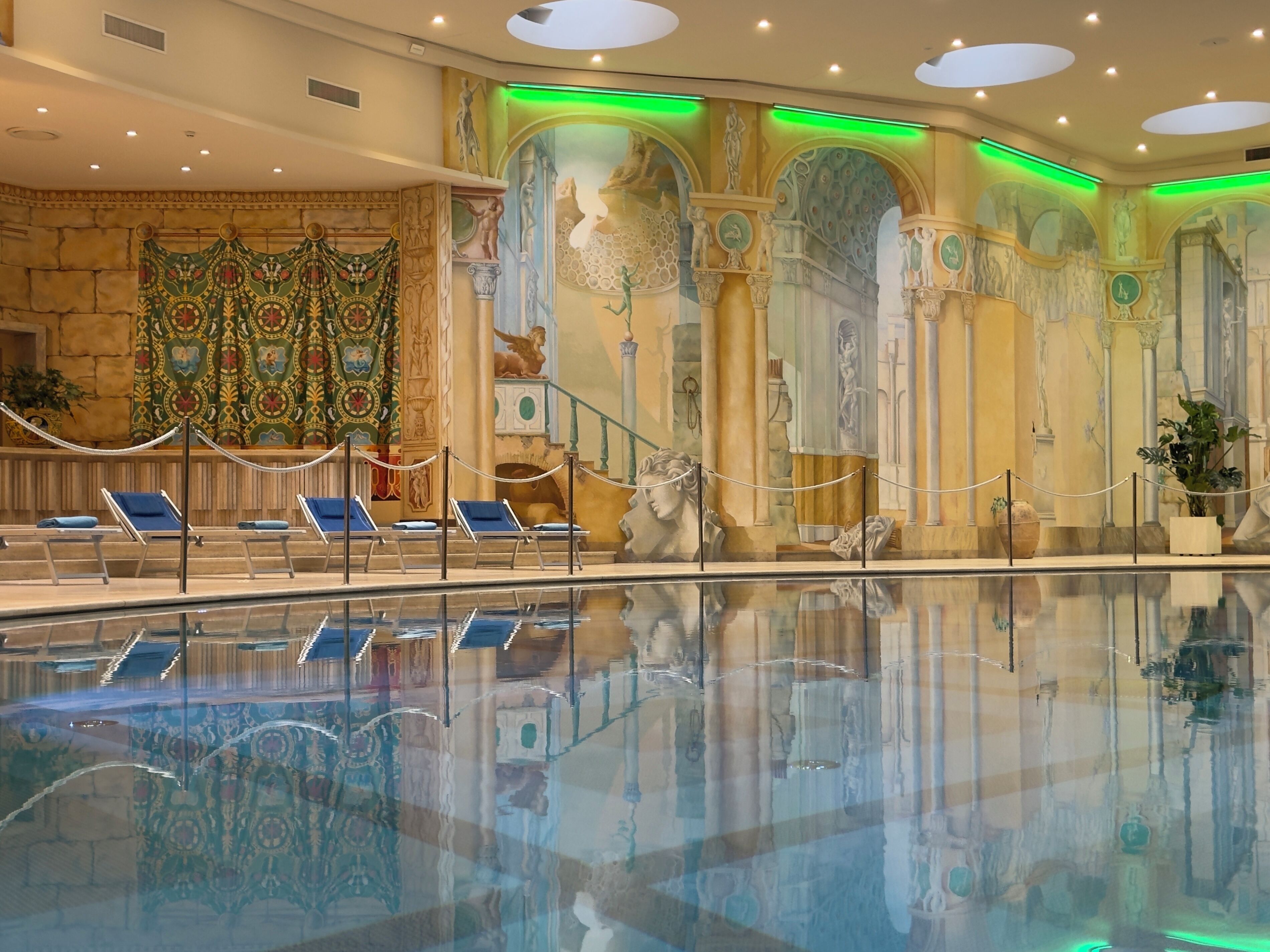 A swimming pool with lounge chairs, a mural with classical figures, and green ambient lighting.