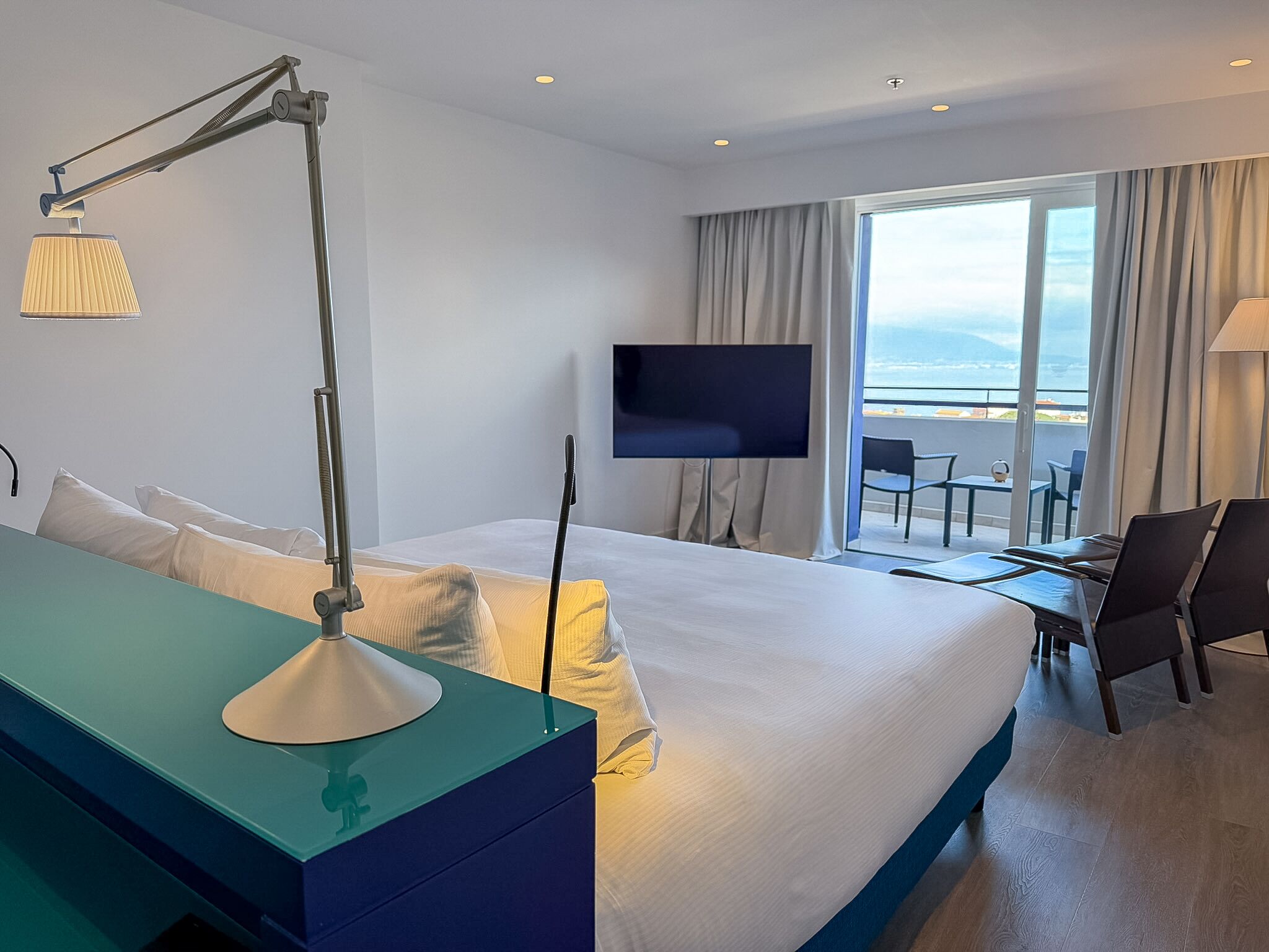 A hotel room with a bed, a desk with a lamp, a TV, and a balcony with a view of the sea.