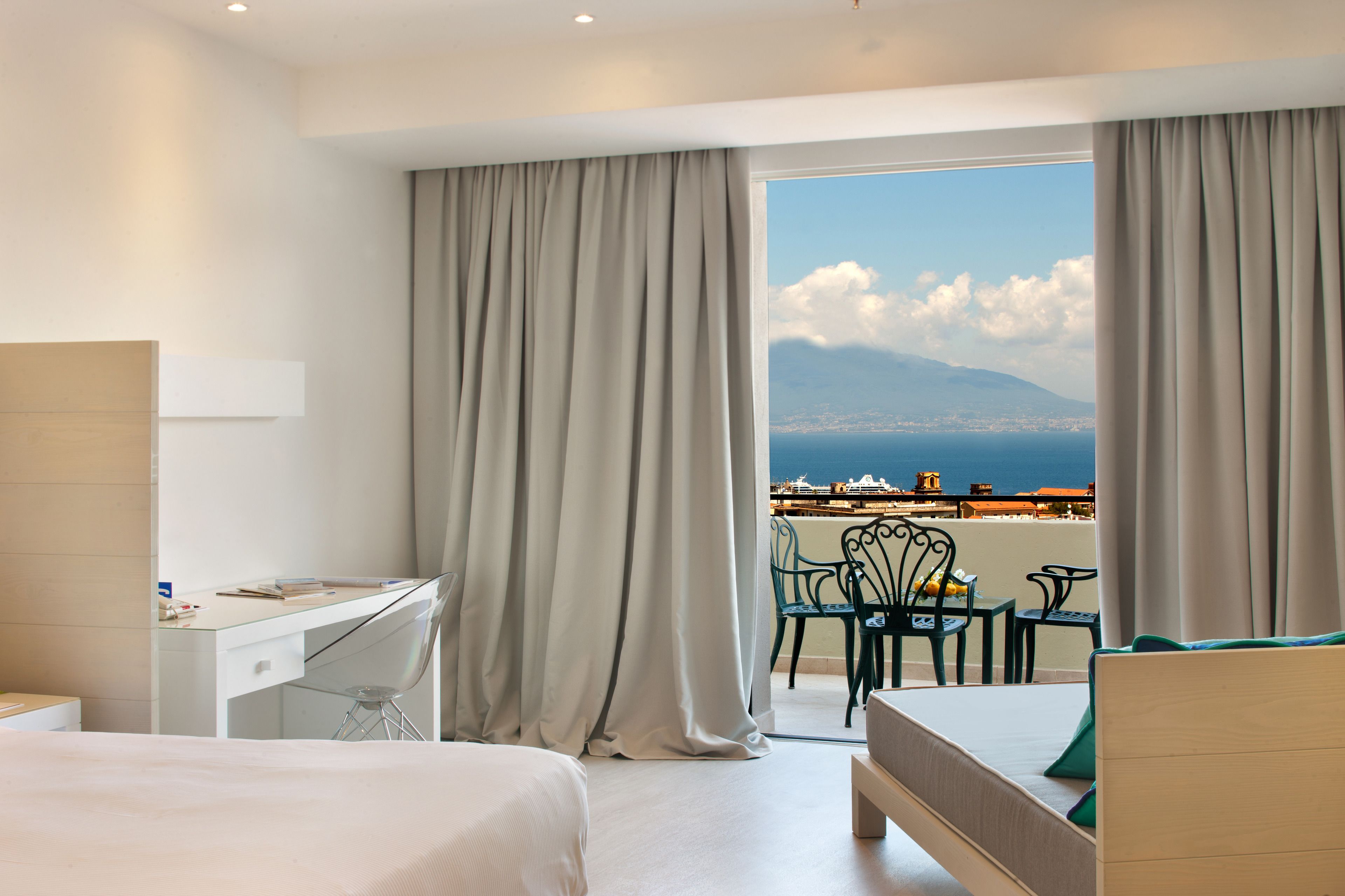 A hotel room with a bed, a desk, and a balcony with a view of the sea and mountains.