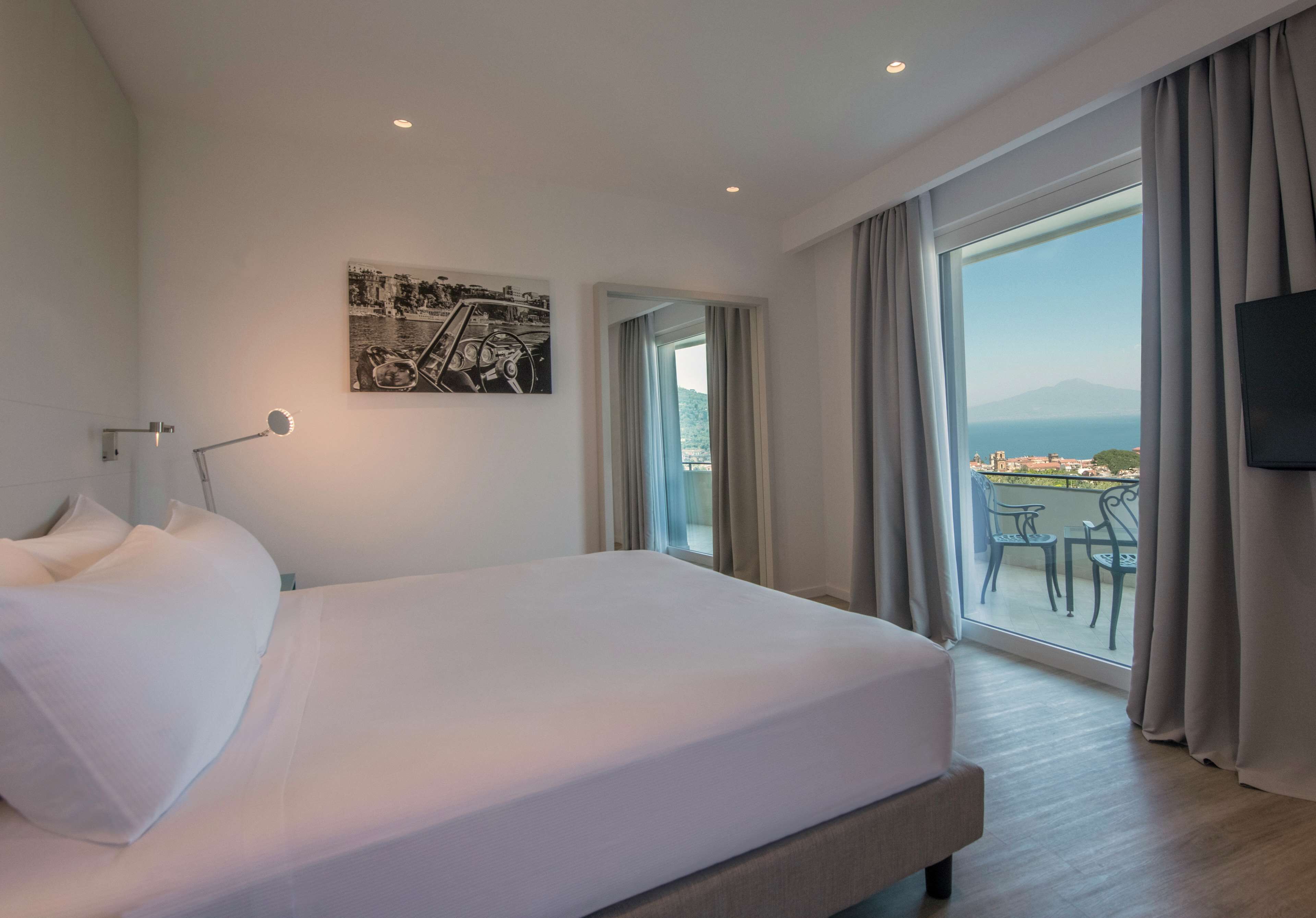 Ein Hotelzimmer mit einem großen Bett, einem Schreibtisch, einem Stuhl, einem Fernseher und einem Balkon mit Blick auf das Meer und die Berge.
