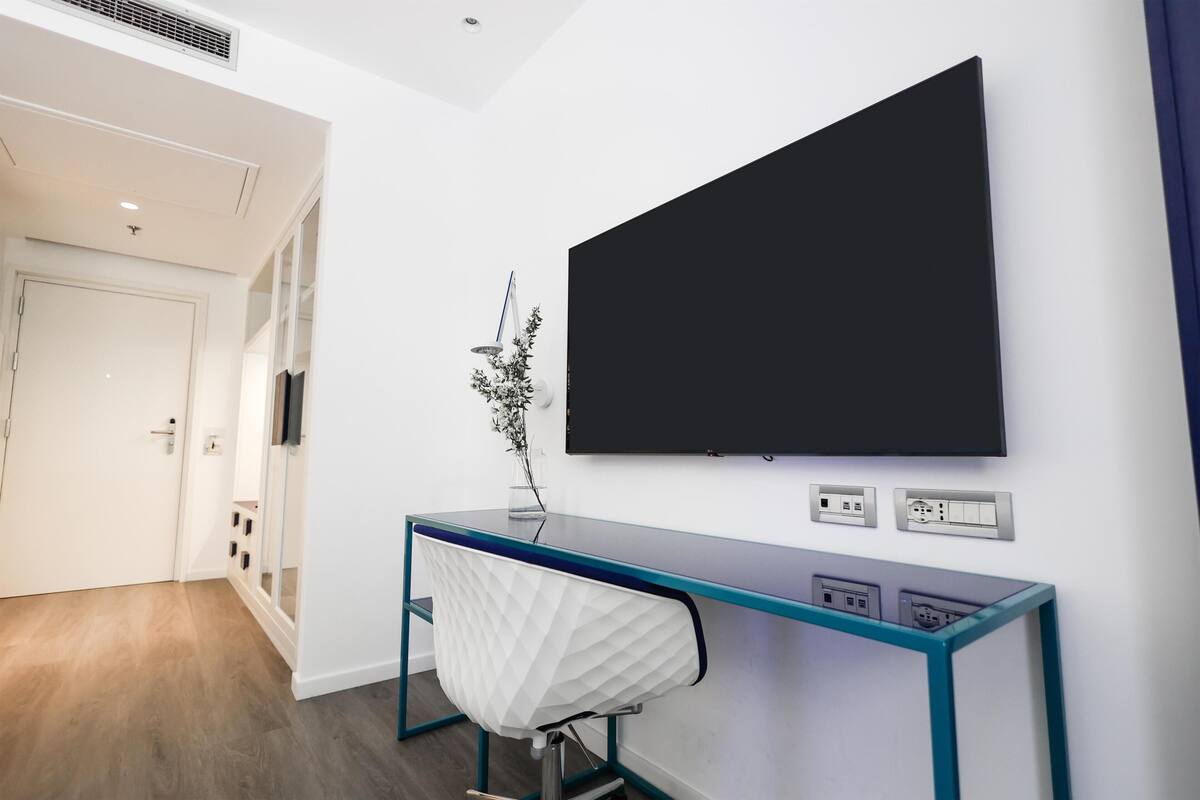 Une chambre moderne dotée d’un téléviseur à écran plat fixé au mur, d’un bureau en verre avec une chaise blanche et d’une porte donnant sur une autre pièce.