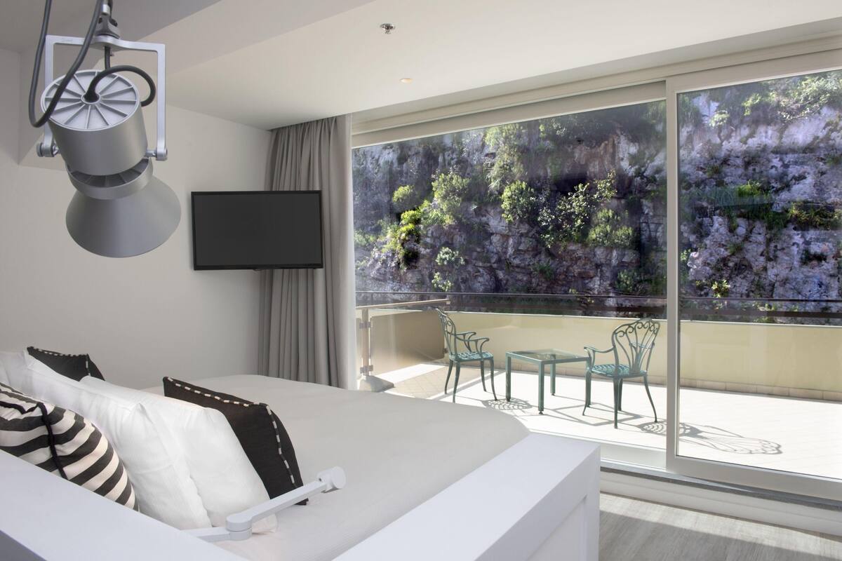 Une chambre d’hôtel moderne dotée d’un grand lit, d’une télévision à écran plat et d’un balcon offrant une vue sur une falaise rocheuse et la verdure.