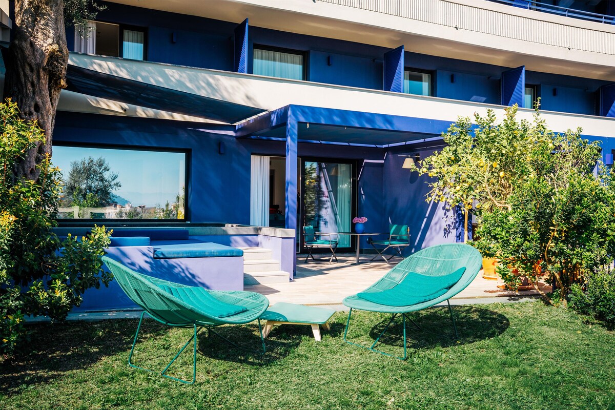 A modern building with a blue exterior, a covered outdoor seating area with green chairs, and a view of the surrounding landscape.