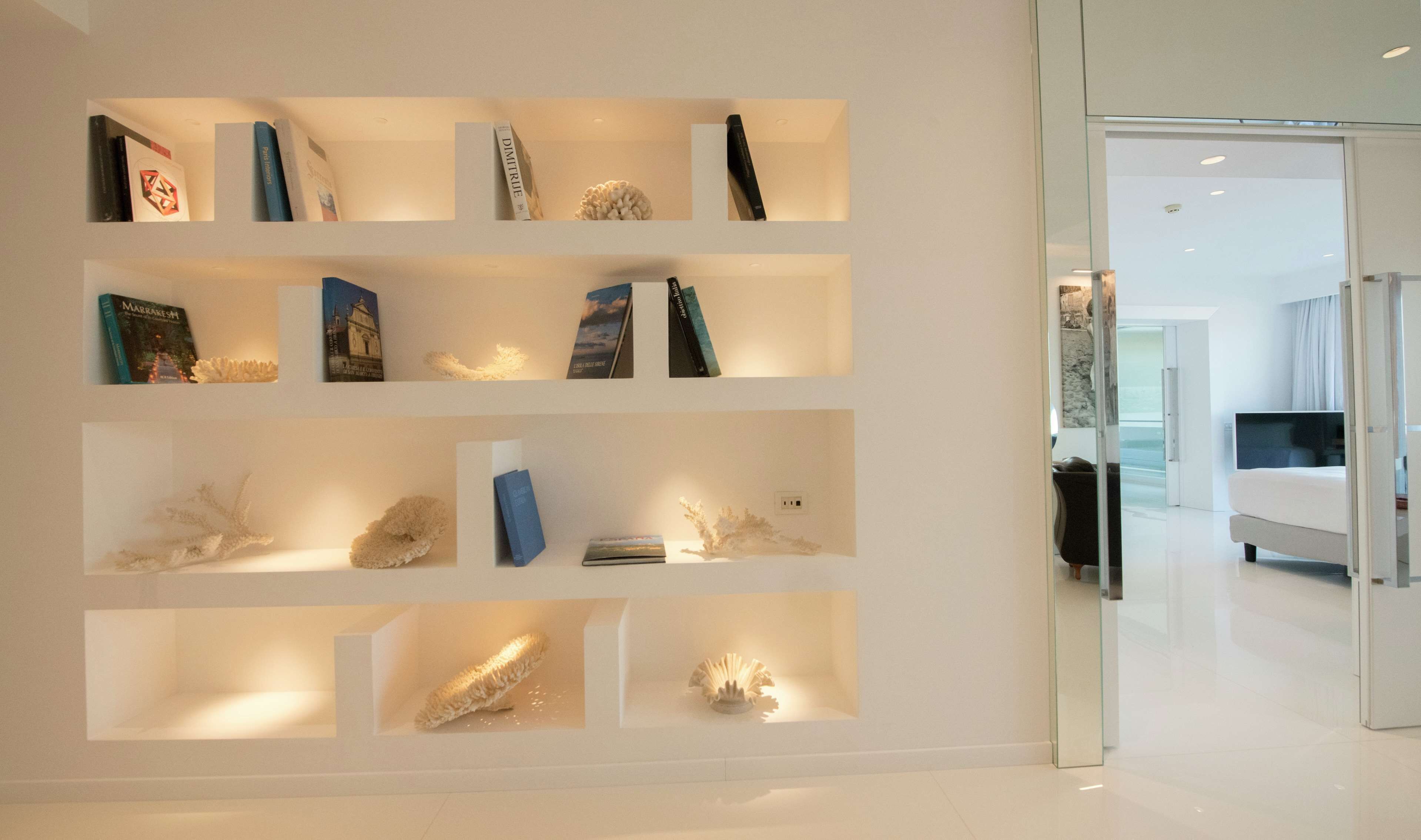 Un intérieur d’hôtel moderne au design minimaliste, avec une bibliothèque murale agrémentée de livres et d’objets décoratifs, une porte en verre donnant sur une chambre équipée d’un lit et d’un téléviseur à écran plat.