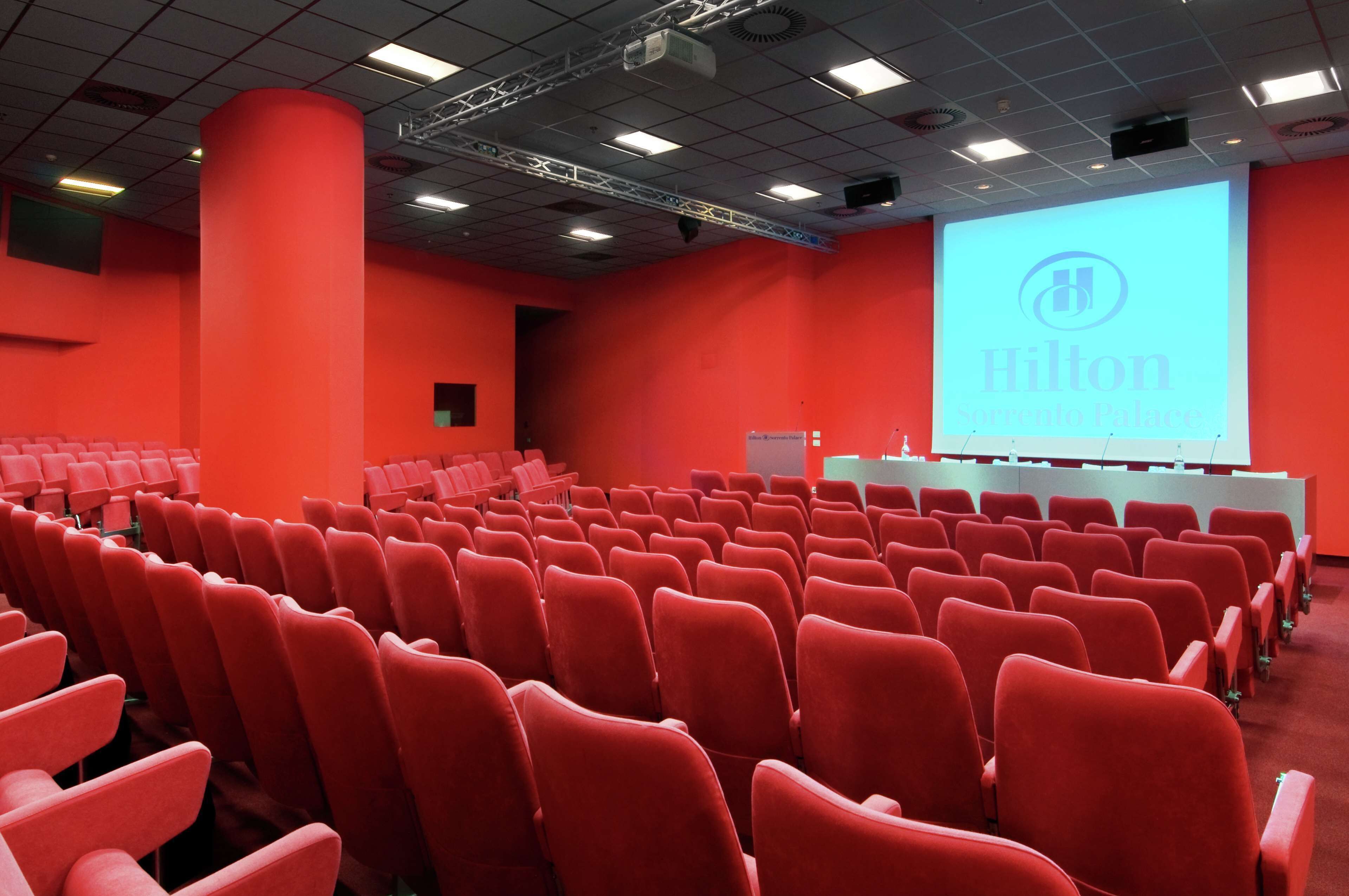 Een congreszaal met rijen rode stoelen, een groot scherm waarop 'Hilton' staat en een podium met een witte tafel.