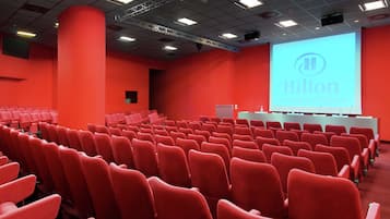 Un salón de conferencias con filas de sillas rojas, una pantalla grande que muestra 'Hilton' y un escenario con una mesa blanca.