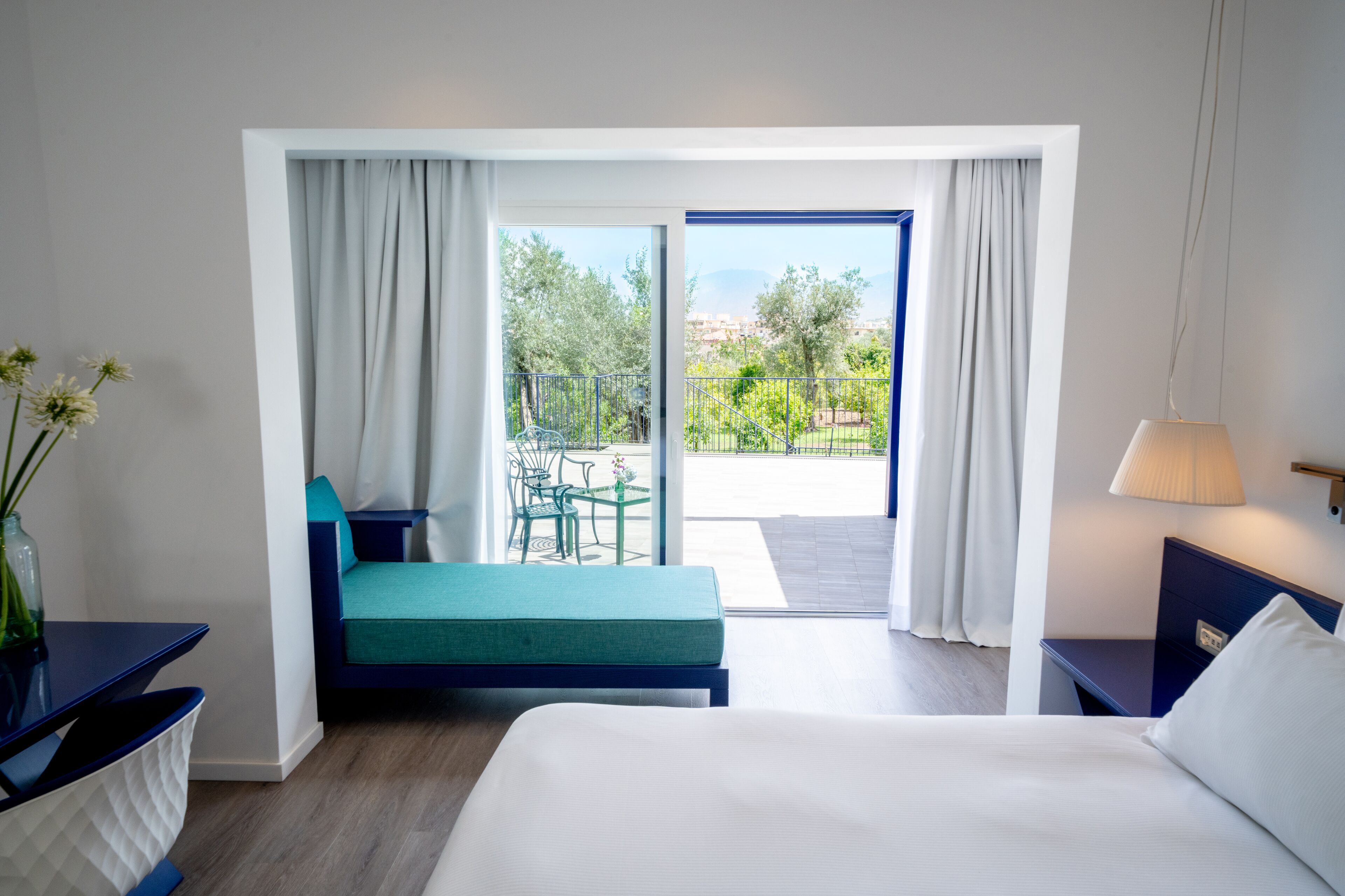 A hotel room with a bed, a small table, a chair, a sofa, a balcony with a view of greenery and buildings, and a lamp.