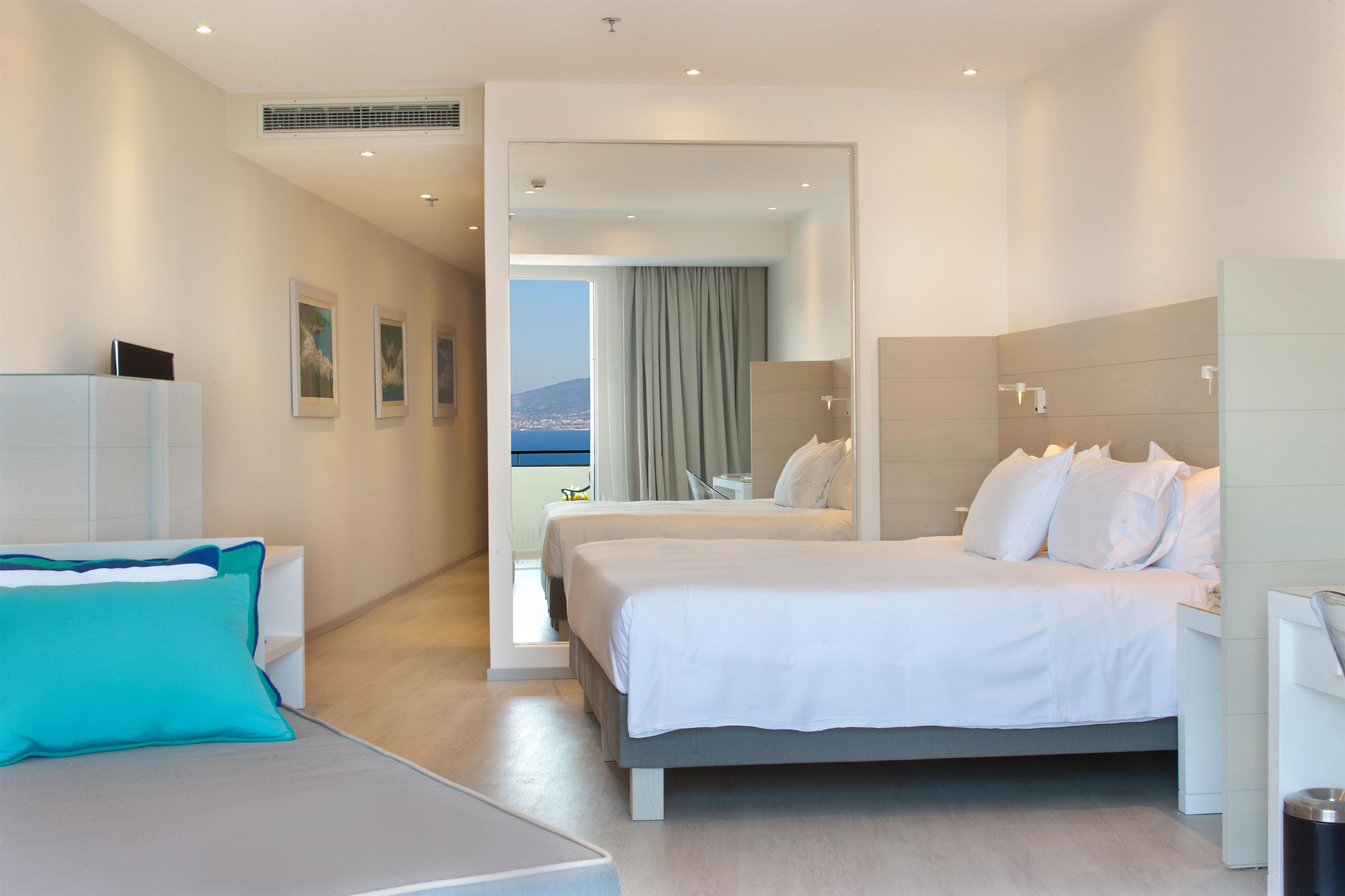 Habitació d'hotel moderna amb un llit gran, zona de seients amb un coixí blau, escriptori i vista al mar per una porta oberta.