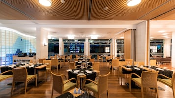 A modern restaurant with multiple dining tables, wooden flooring, and a ceiling with linear lighting fixtures.