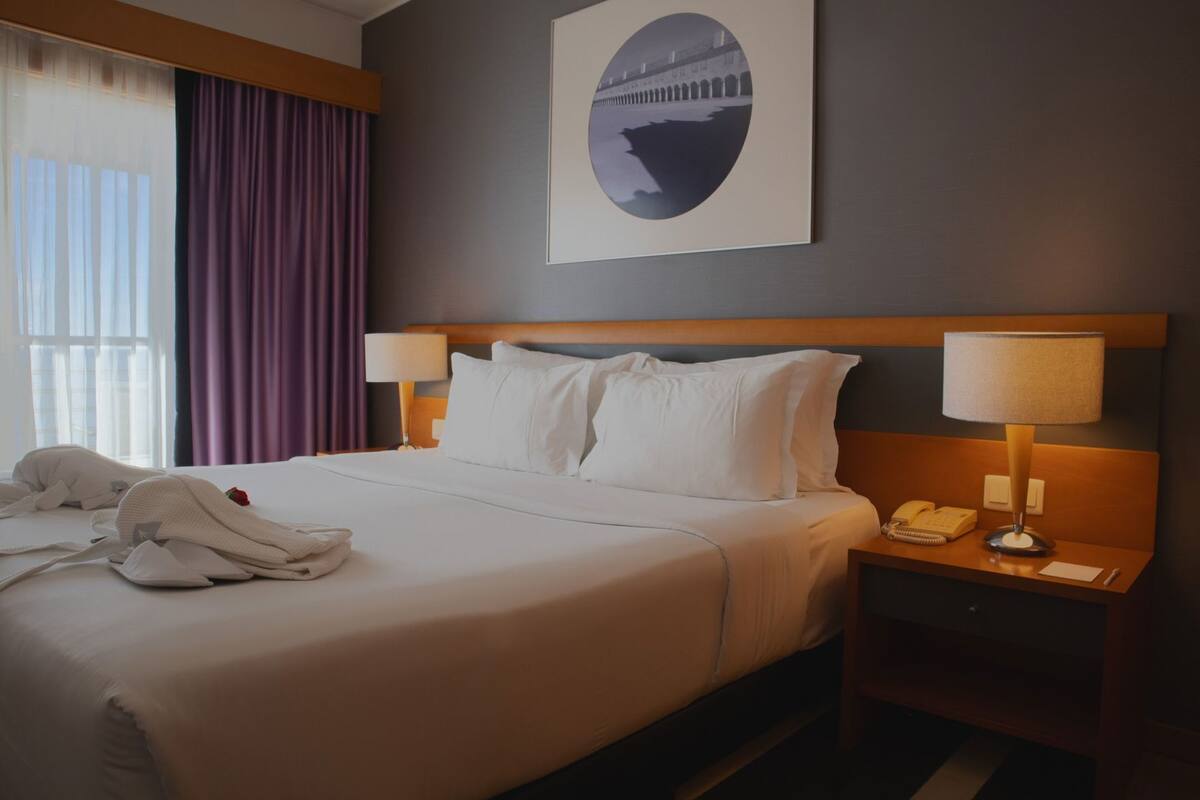 Une chambre d’hôtel avec un grand lit, deux tables de chevet avec des lampes, un tableau encadré au mur et une fenêtre avec des rideaux.