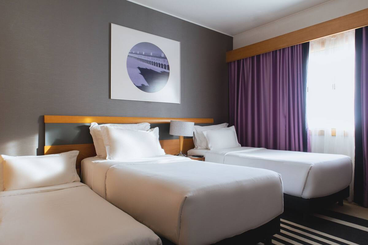 Une chambre d’hôtel avec deux lits, une tête de lit, un tableau encadré au mur et une fenêtre avec des rideaux violets.