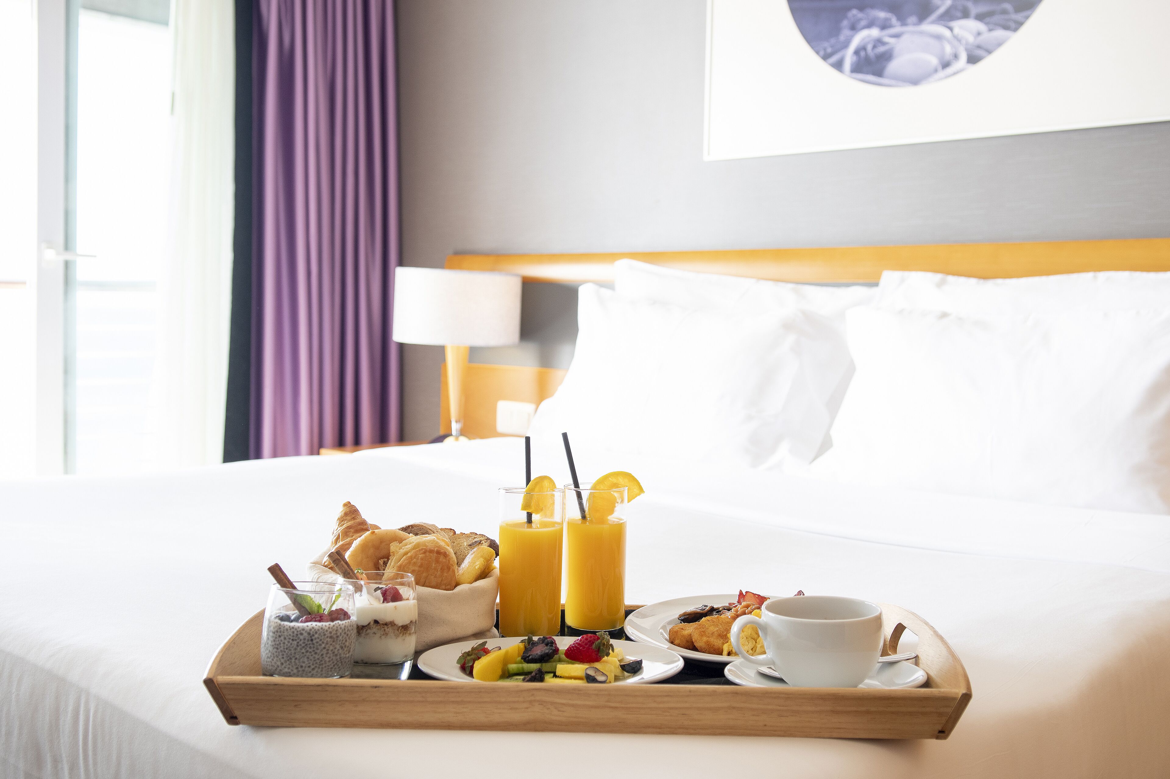 A neatly made breakfast tray on a bed with croissants, fruit, yogurt, and juice glasses.