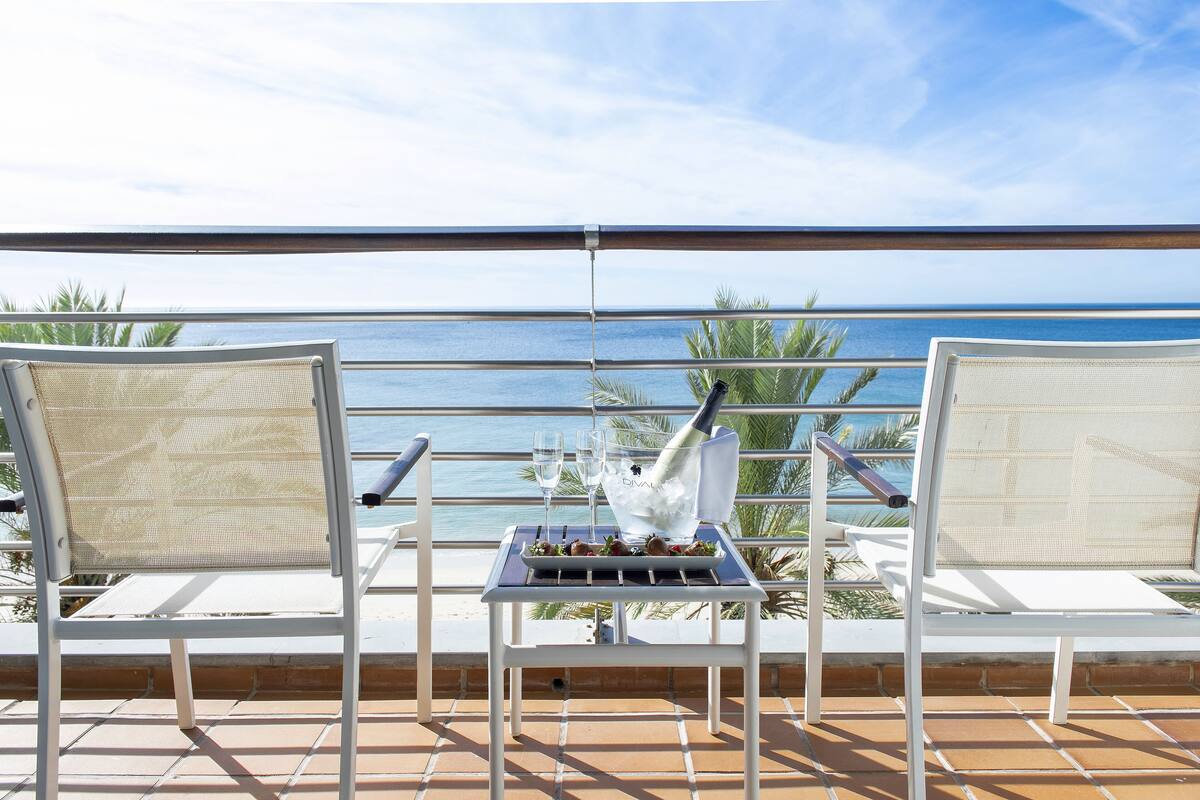 Un balcon avec deux chaises blanches, une table sur laquelle se trouvent une bouteille de champagne, des verres et un plateau de nourriture. La vue comprend la mer et des palmiers.