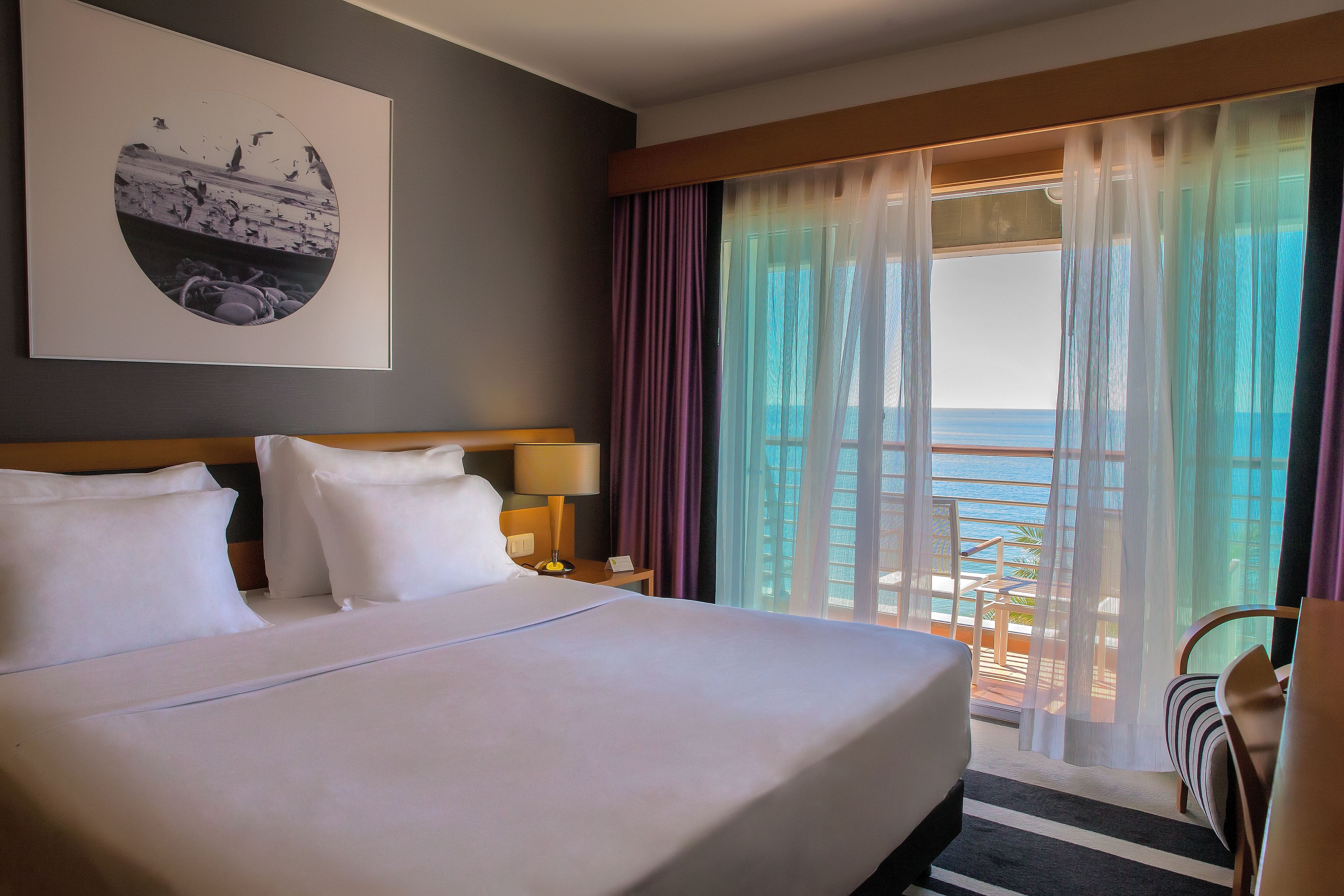 Habitación de hotel con una cama grande, una mesita de noche, una lámpara, vista al océano a través de cortinas translúcidas y un cuadro en la pared.