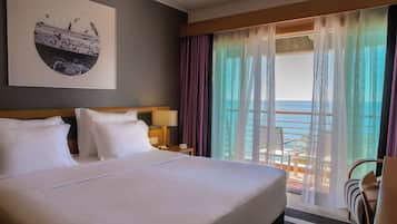 Habitación de hotel con una cama grande, una mesita de noche, una lámpara, vista al océano a través de cortinas translúcidas y un cuadro en la pared.