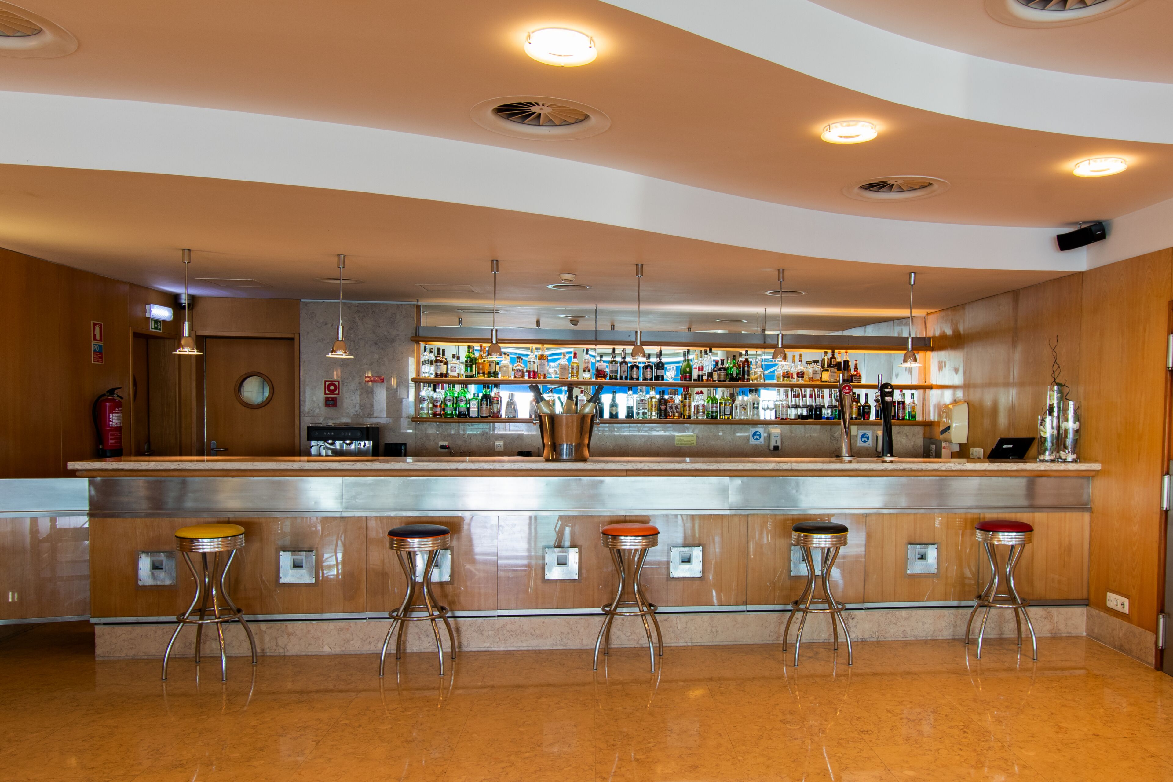 Een bar met een gebogen toonbank, vier krukken en diverse flessen en glazen op planken.